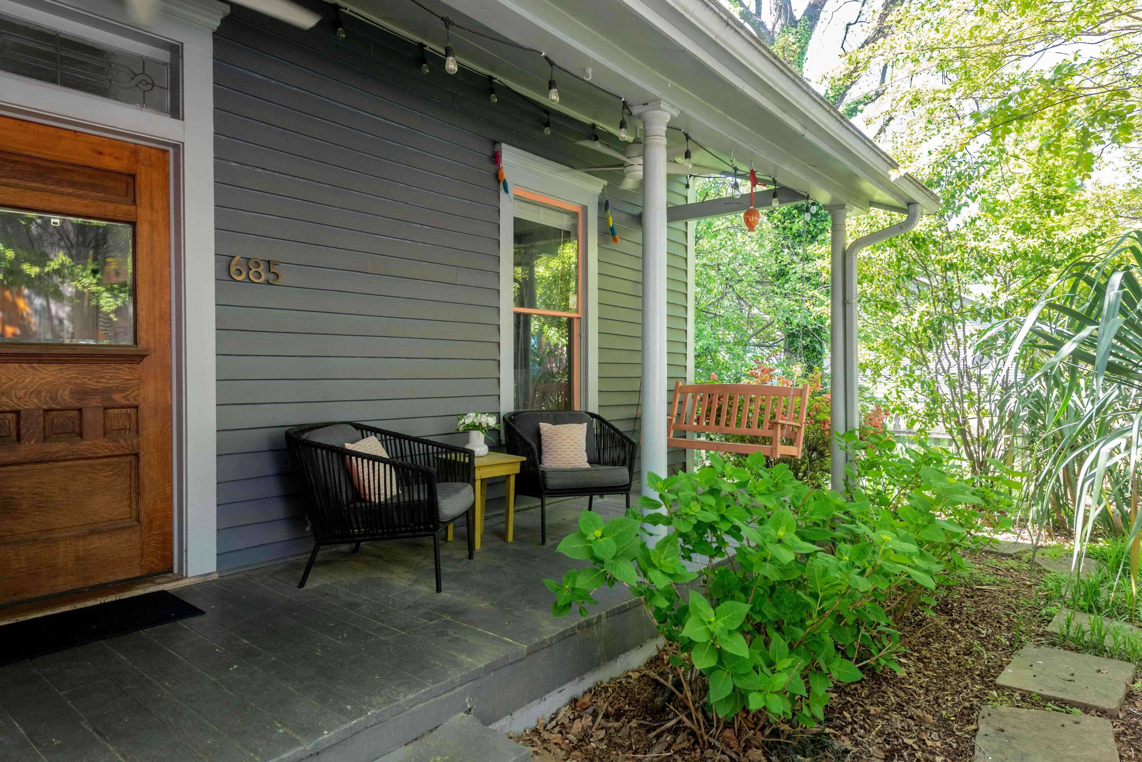The image shows a cozy front porch with two black chairs, a small table, and a swing bench surrounded by greenery.