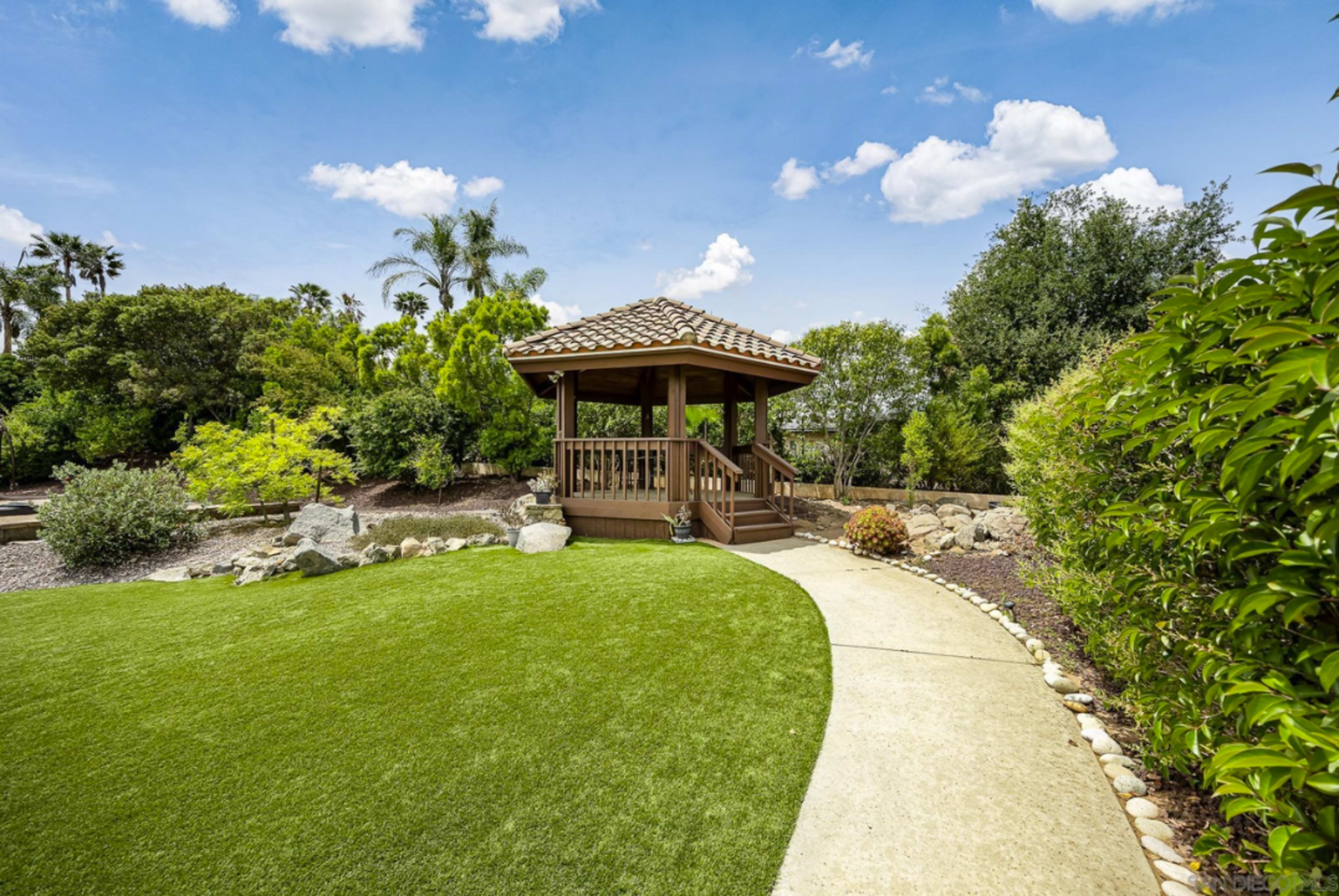 A gazebo sits on a manicured lawn with a concrete pathway leading to it, surrounded by lush greenery and rocks.