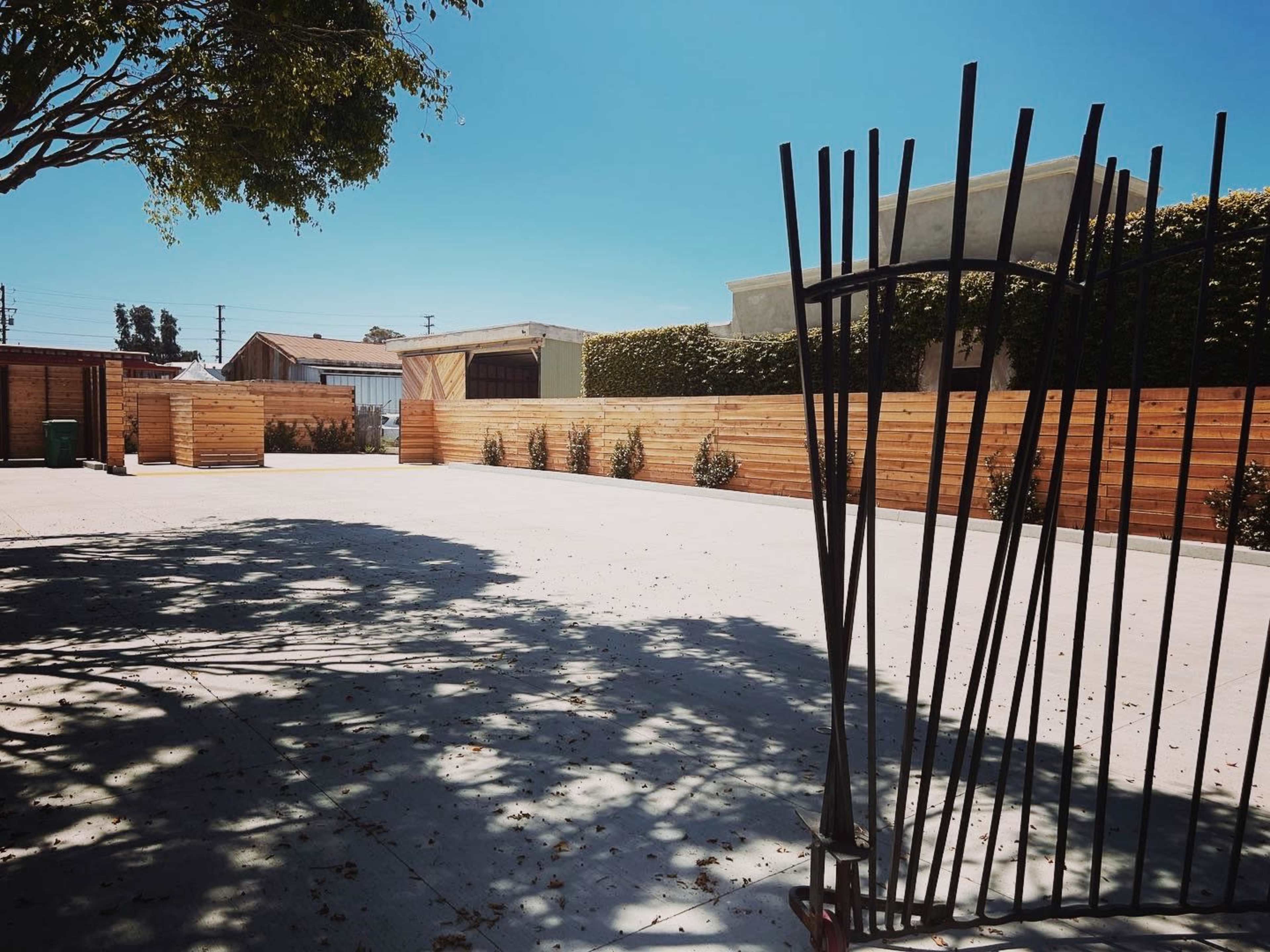 A cleared outdoor space with a metal gate and wooden fencing, surrounded by trees and buildings.