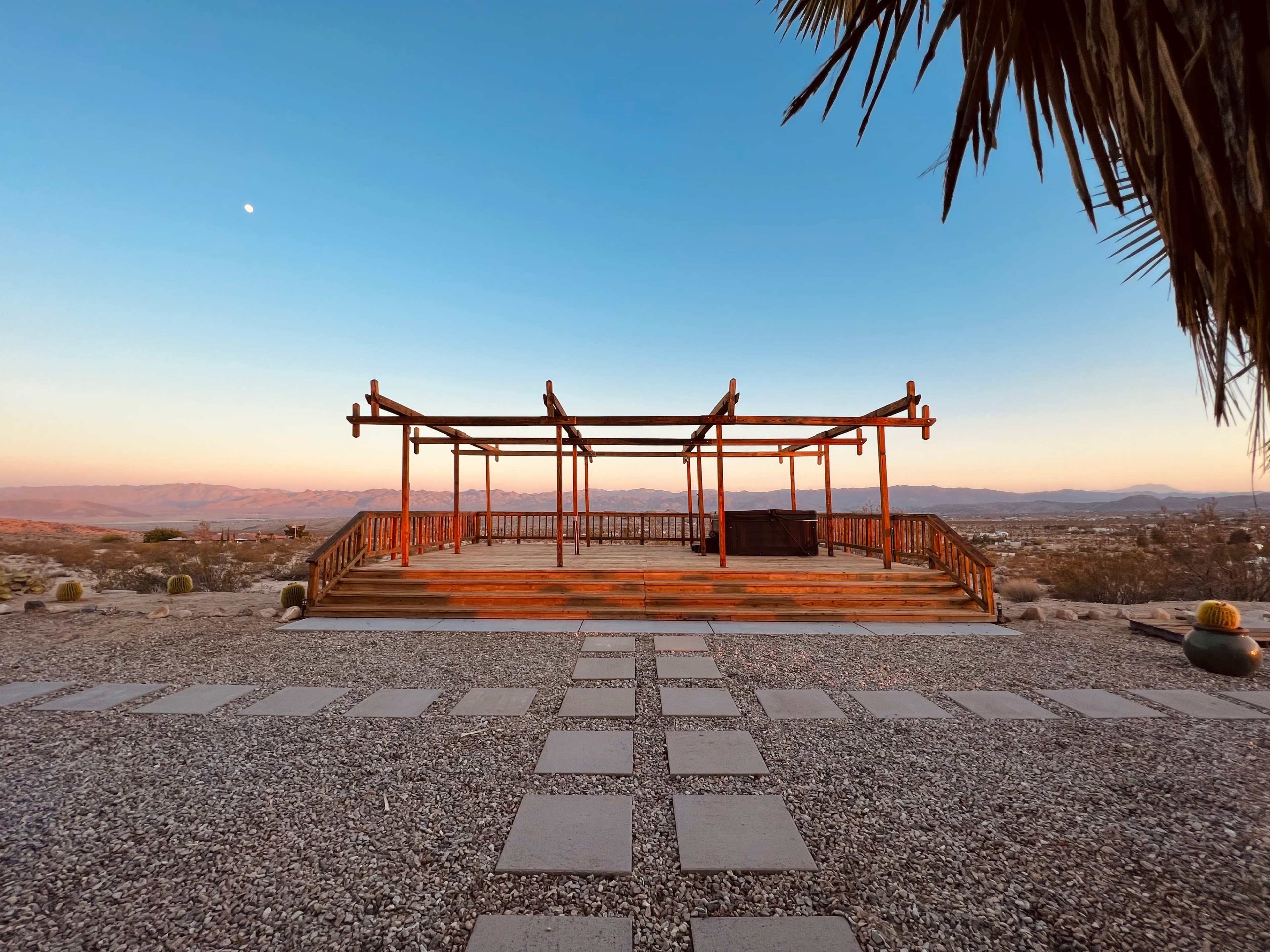 An open wooden framework stands on a gravel surface, overlooking a desert landscape at dusk with a crescent moon visible in the sky.