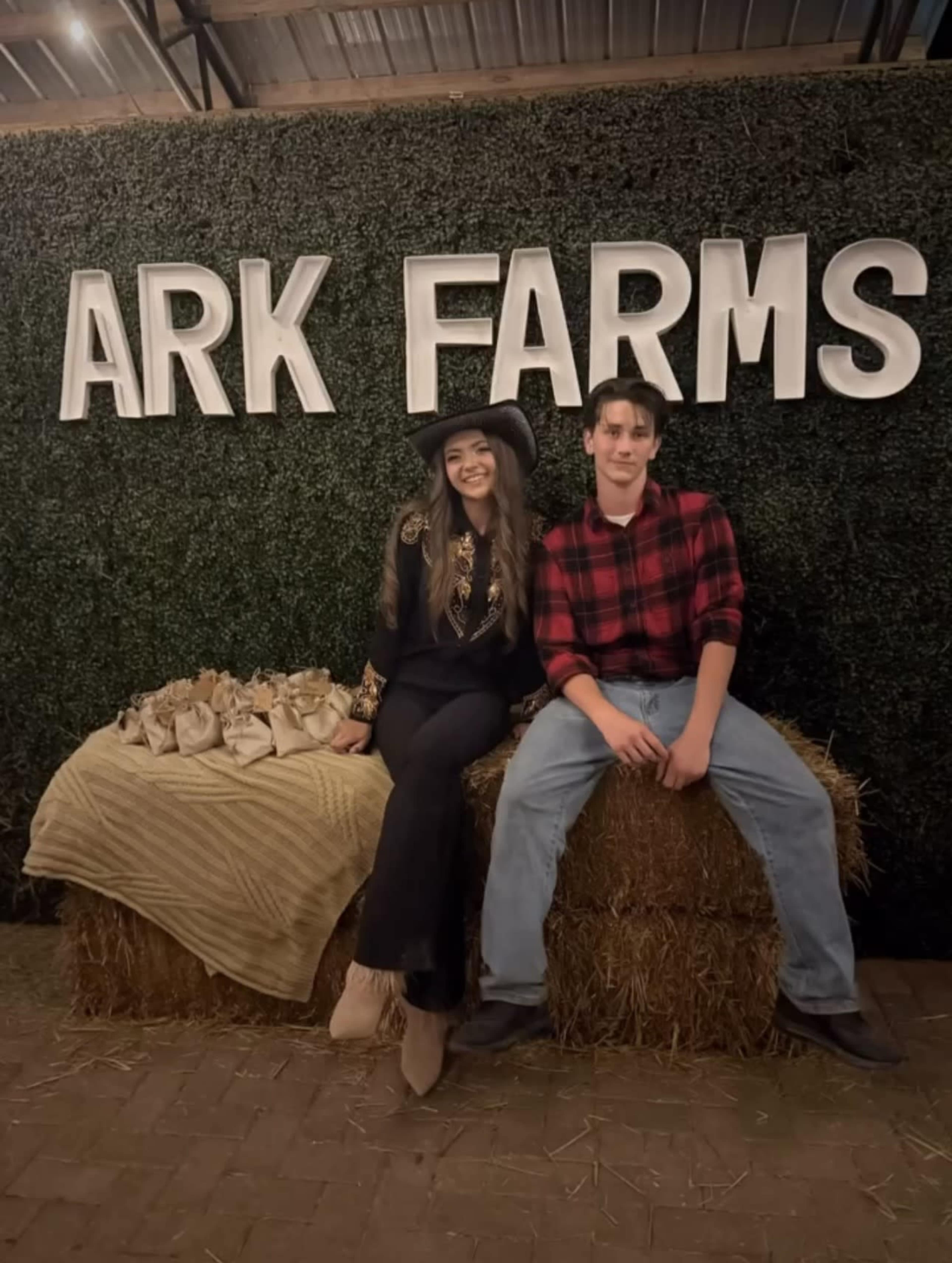 Two people are sitting on a hay bale in front of a decorative wall with the words "ARK FARMS" displayed prominently.