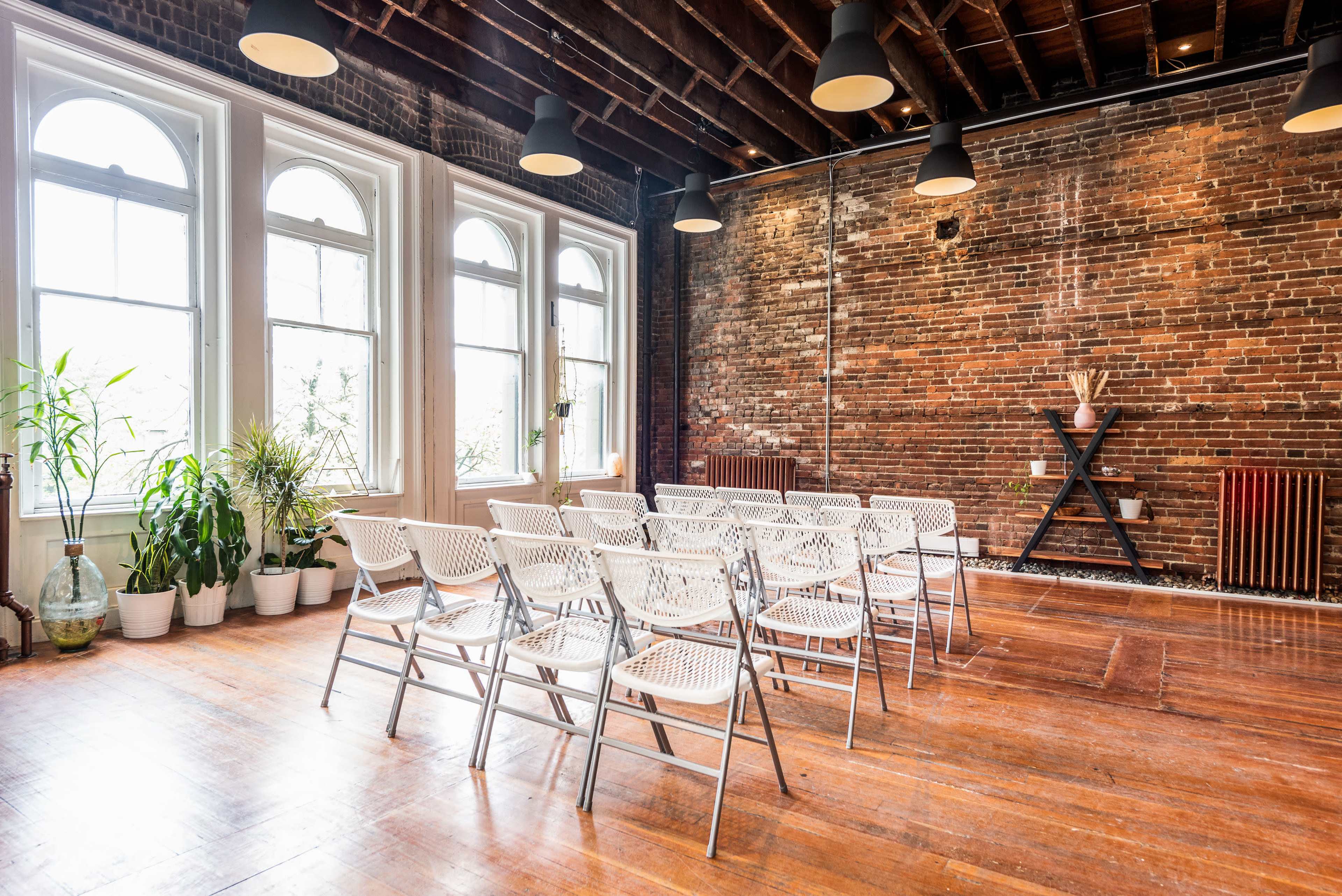 A room with exposed brick walls, wooden floors, and a setup of white folding chairs arranged for an event, with large windows allowing natural light and potted plants in the corners.