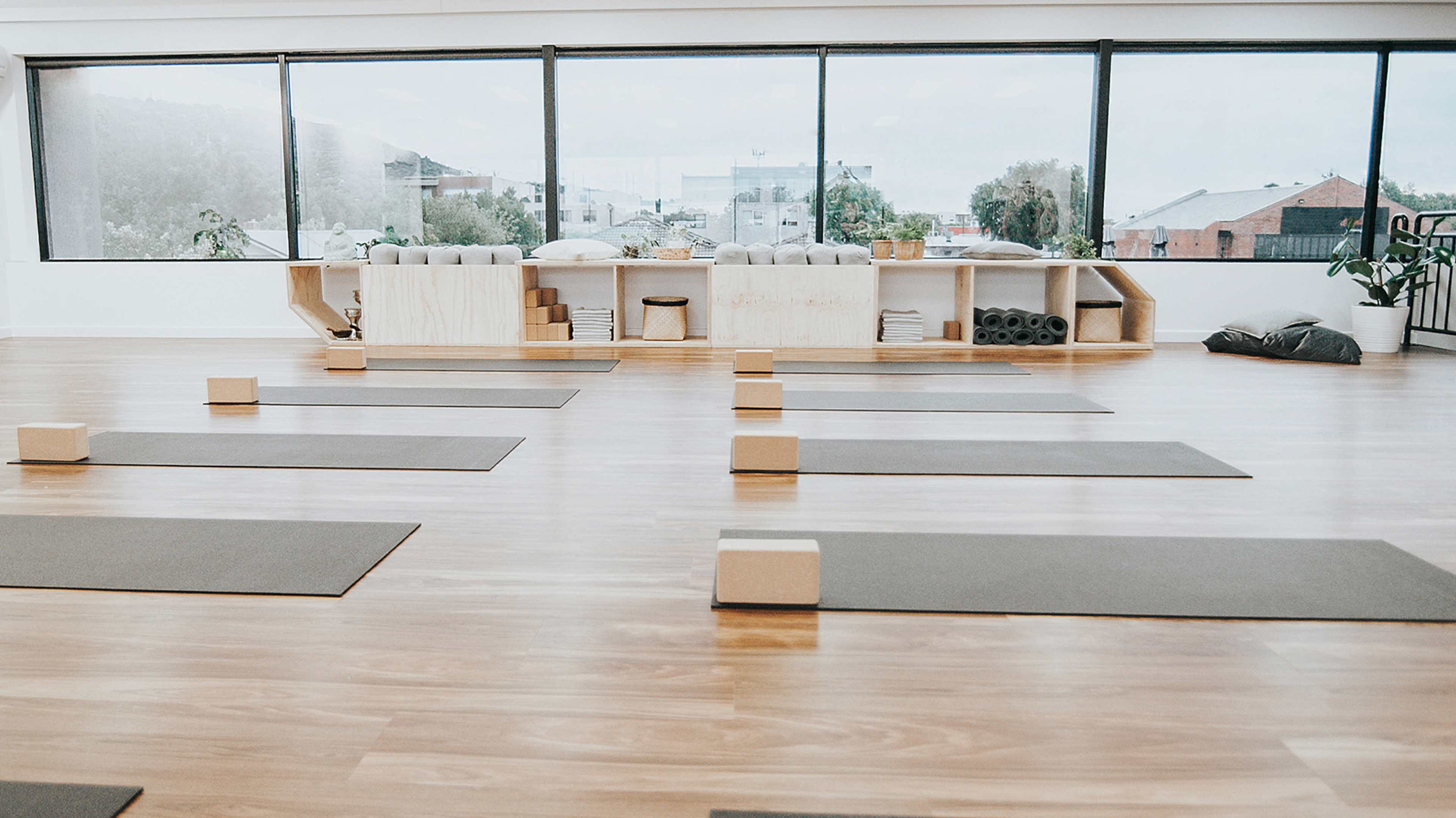 A spacious yoga studio with several yoga mats arranged on a wooden floor, blocks placed on the mats, and a seating area with storage shelves in the background.
