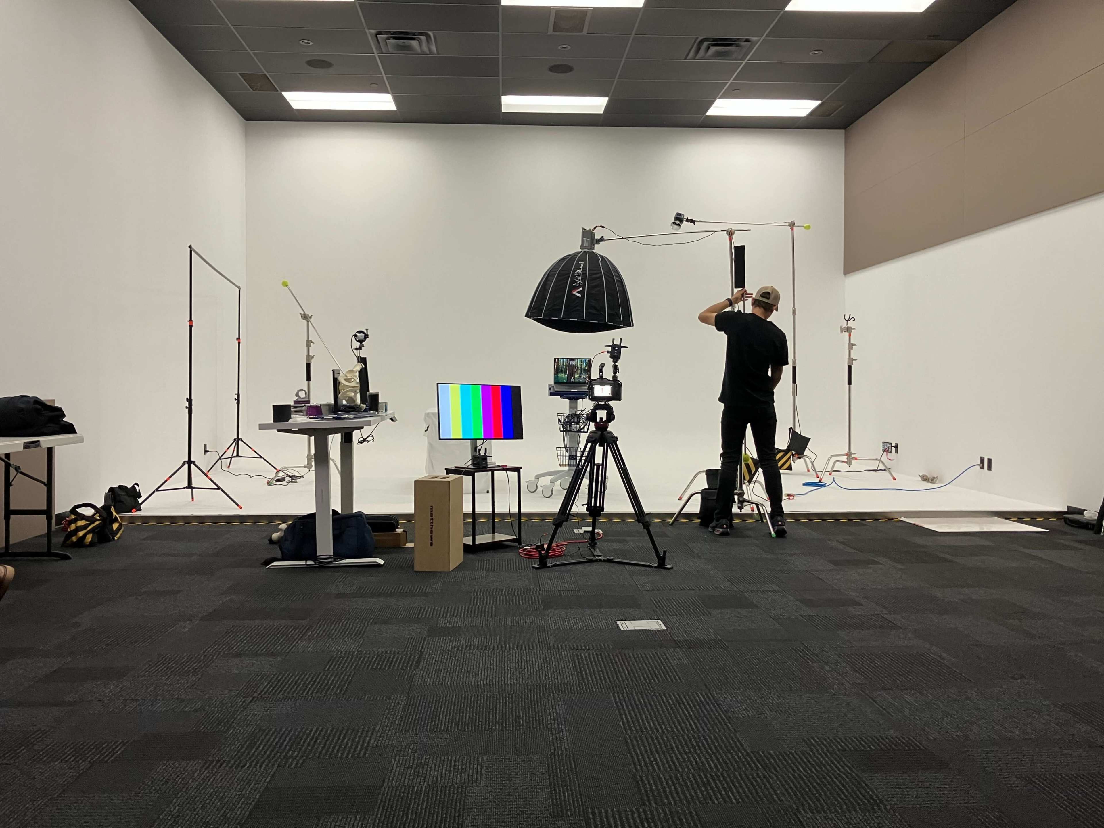 A studio setup features a camera, lighting equipment, a monitor displaying color bars, and a person adjusting equipment in a blank, white-walled space.