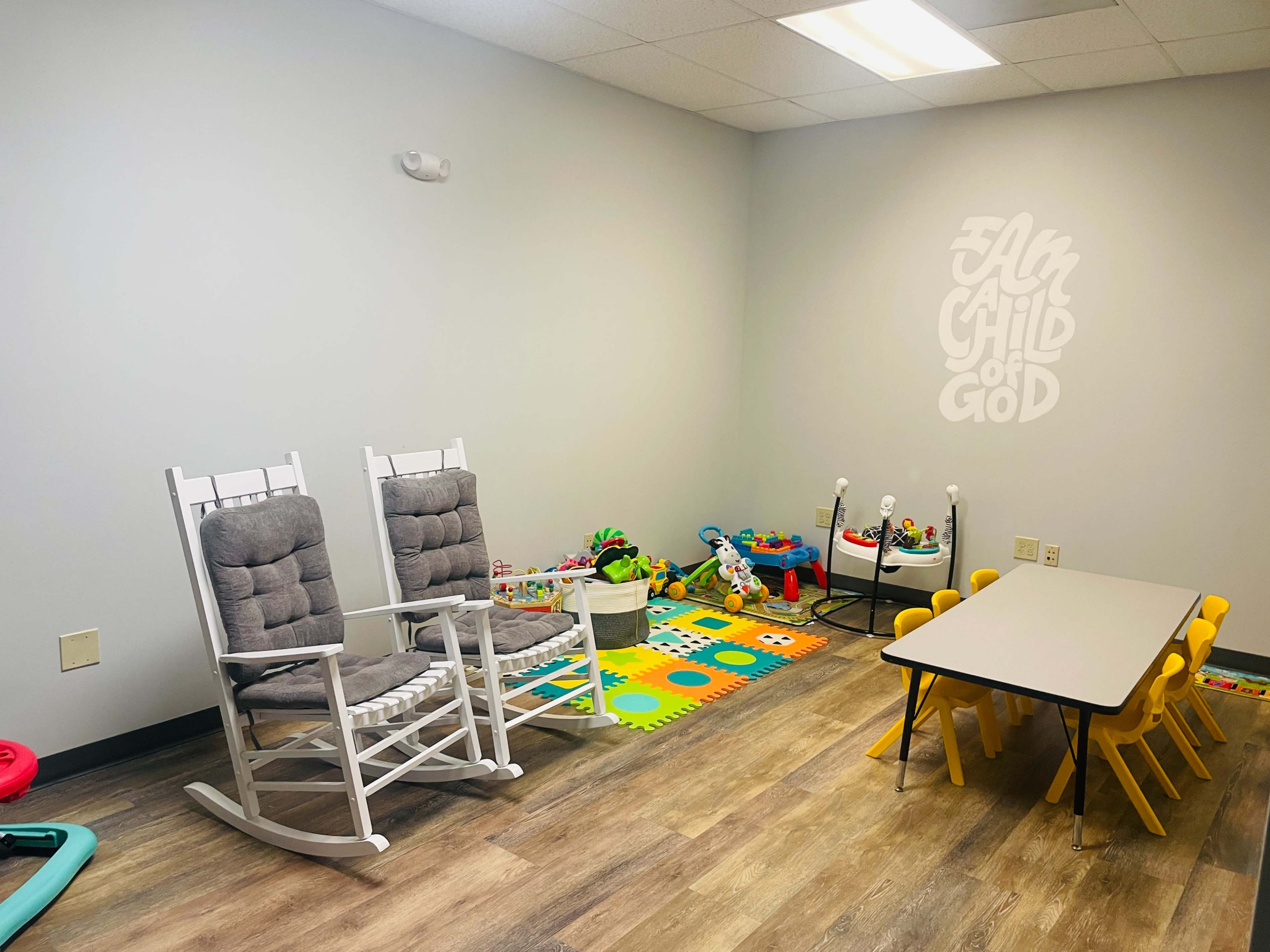 A children's playroom featuring two rocking chairs, a colorful play area with toys, and a table surrounded by small yellow chairs.