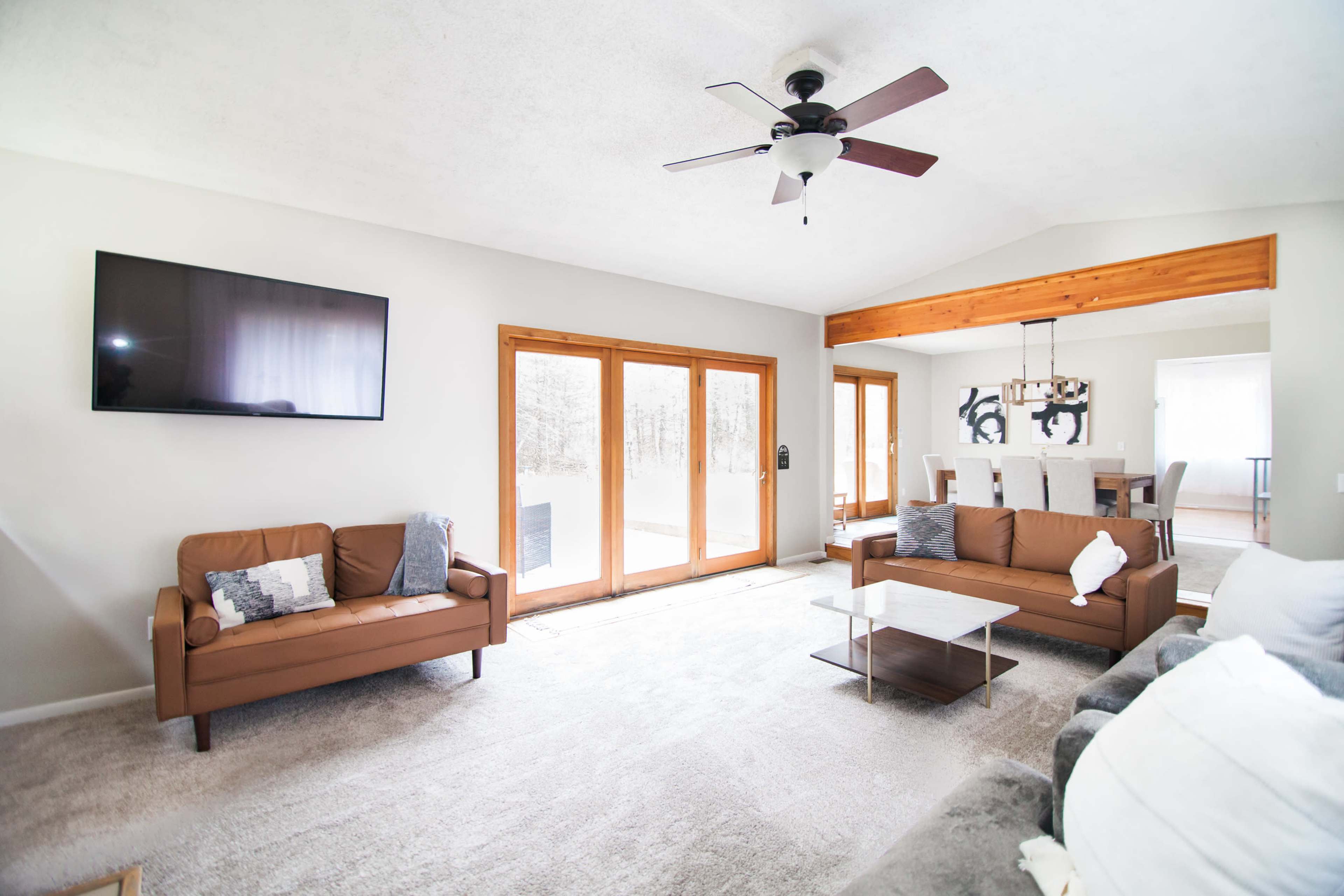 A spacious living room features two brown sofas, a glass coffee table, a wall-mounted television, and large windows that provide natural light.