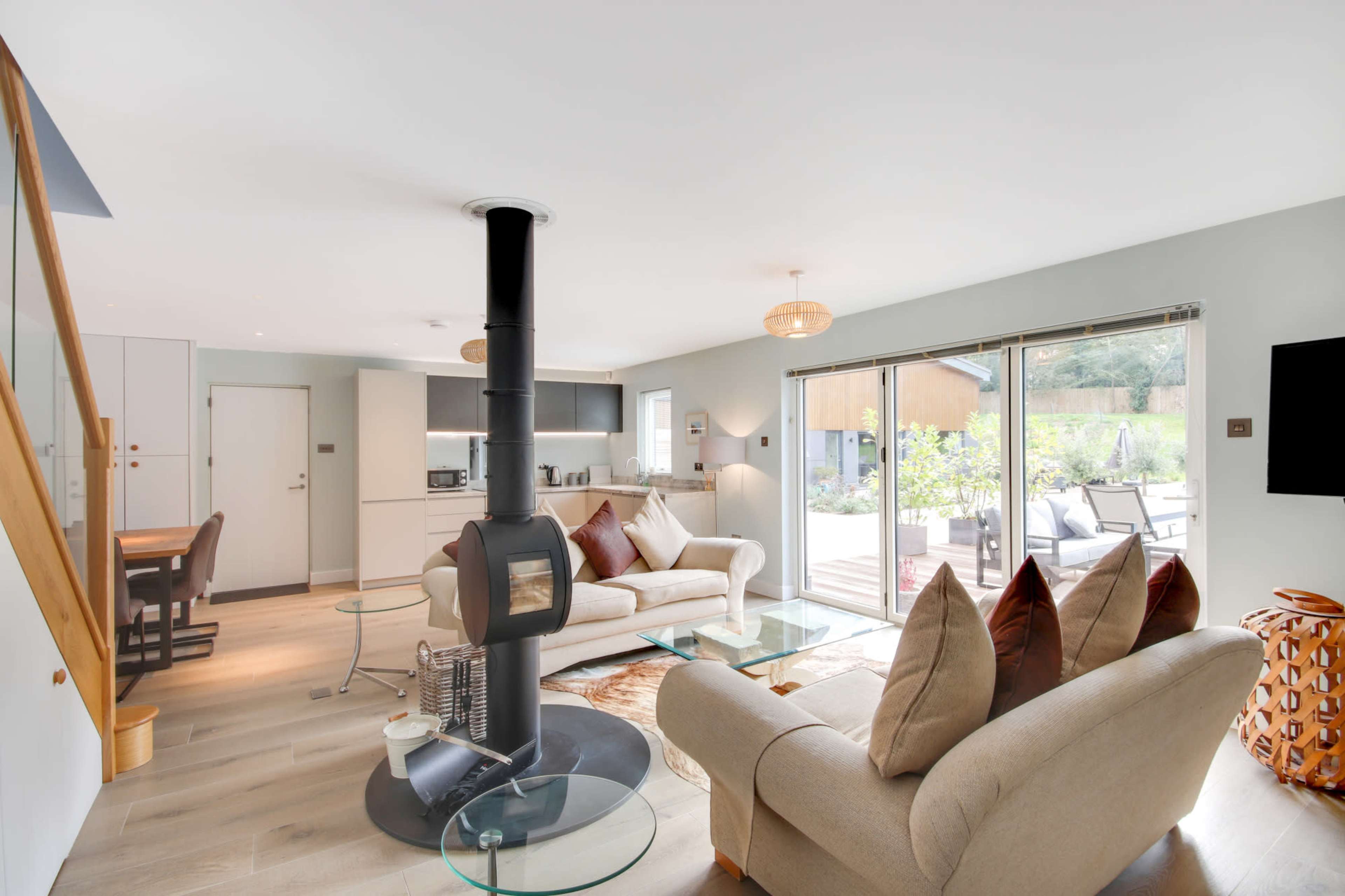 The image shows a modern living room with a wood-burning stove, a glass coffee table, and large sliding doors that open to an outdoor patio.