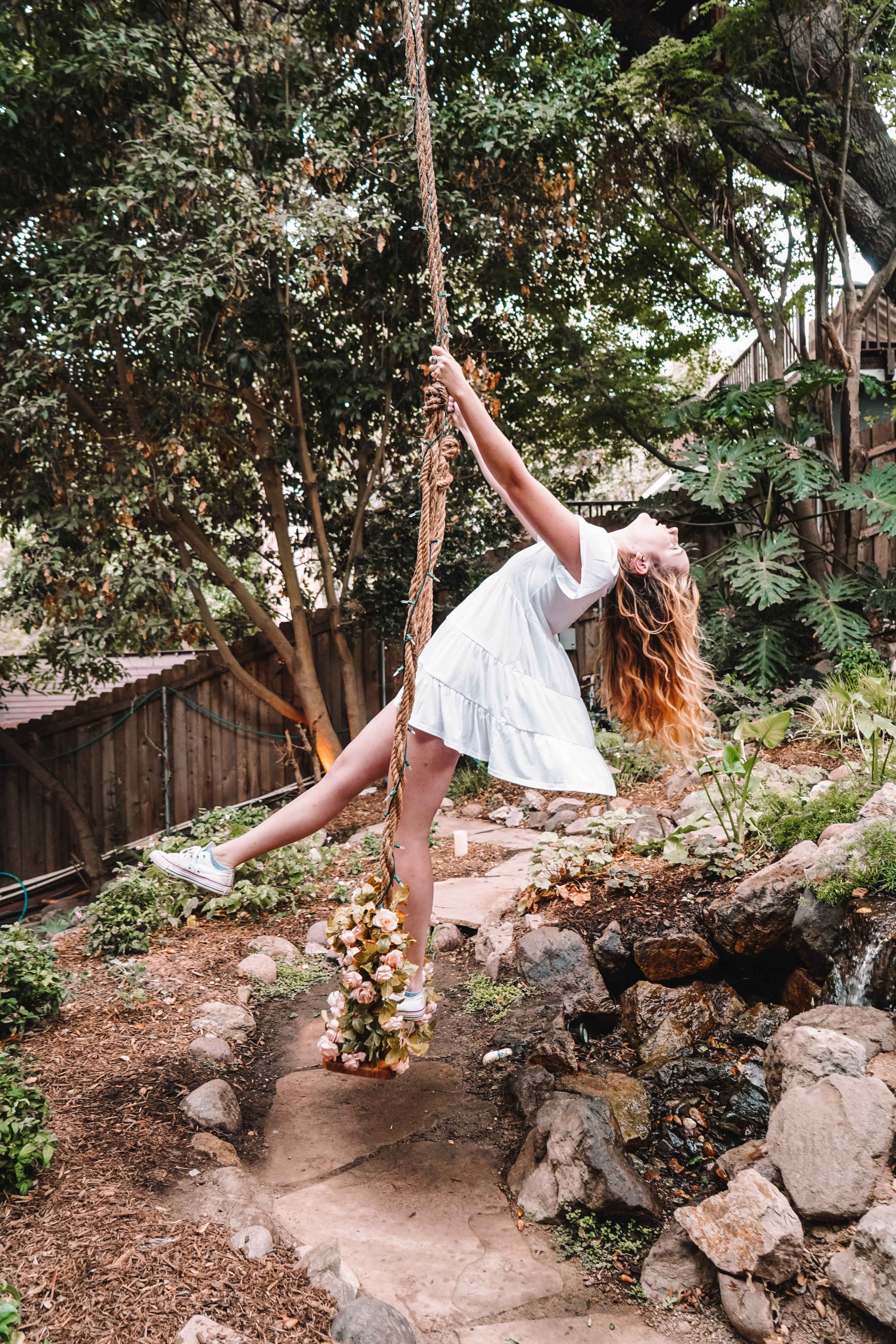 A person swings on a rope tied to a tree in a garden, surrounded by greenery and rocks.
