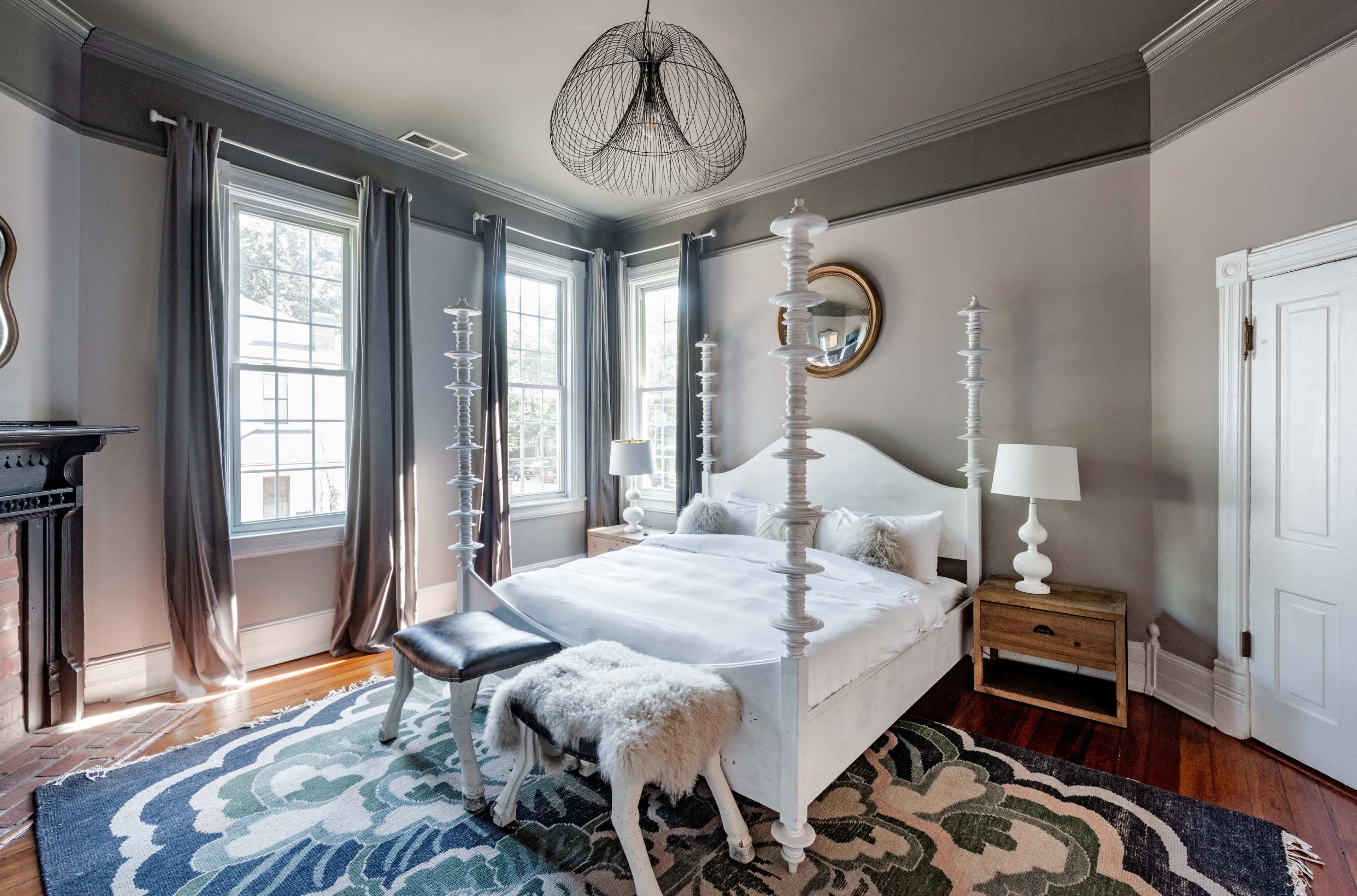 The image shows a well-lit bedroom with a white bed, two windows, and a decorative area rug.