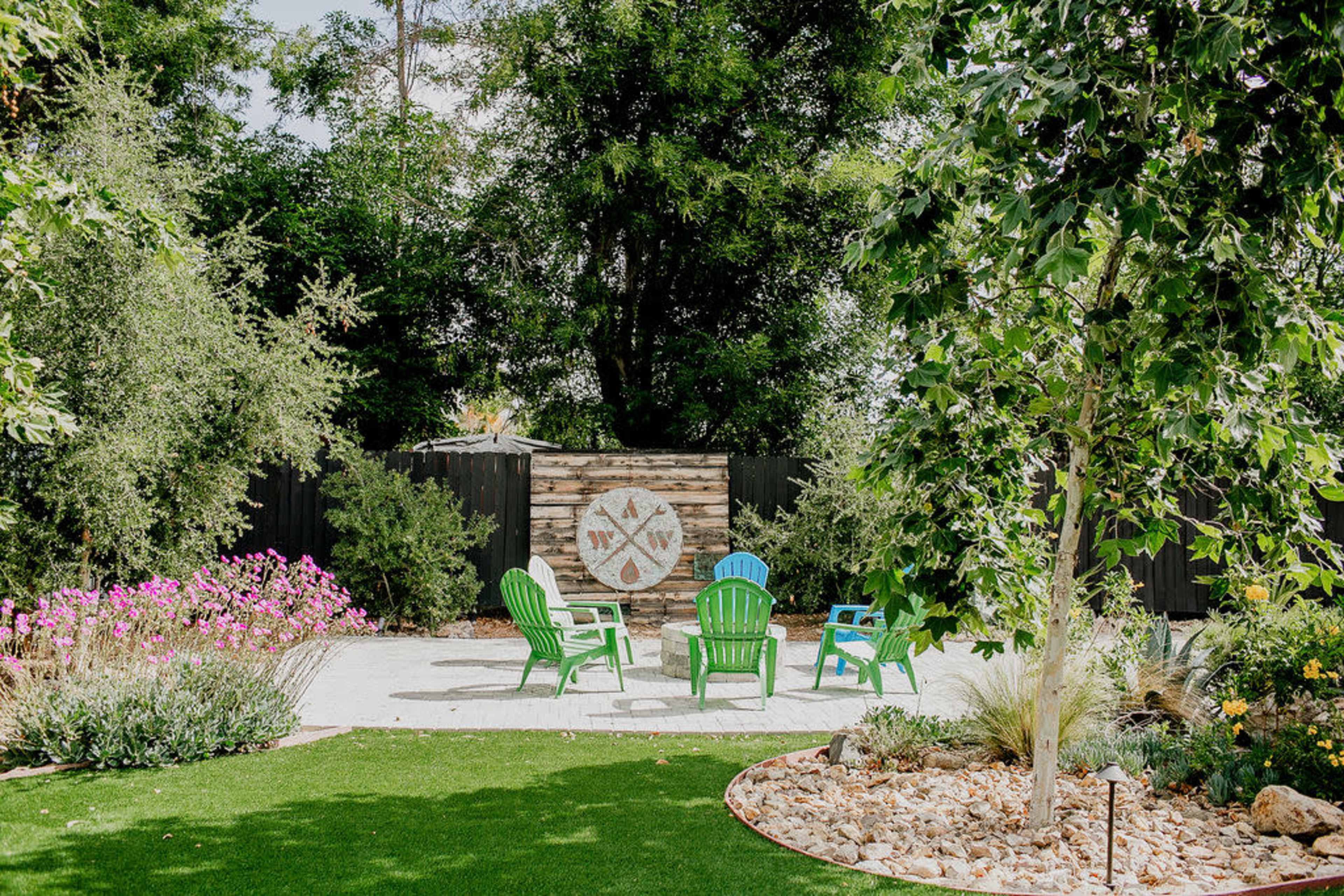 A landscaped garden features green chairs arranged around a stone pathway, with a wooden backdrop and flowering plants.