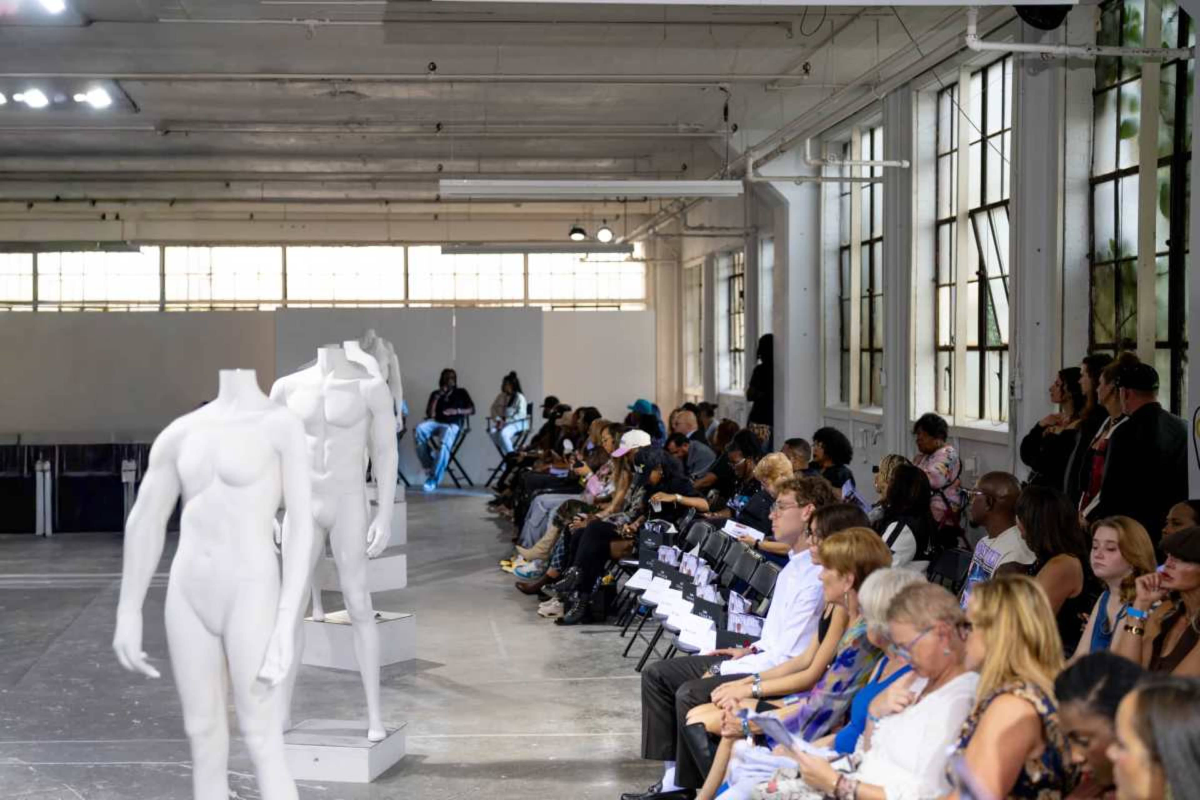 The image shows a fashion event with a row of empty chairs facing white mannequins on display in a spacious, brightly lit venue filled with an audience.