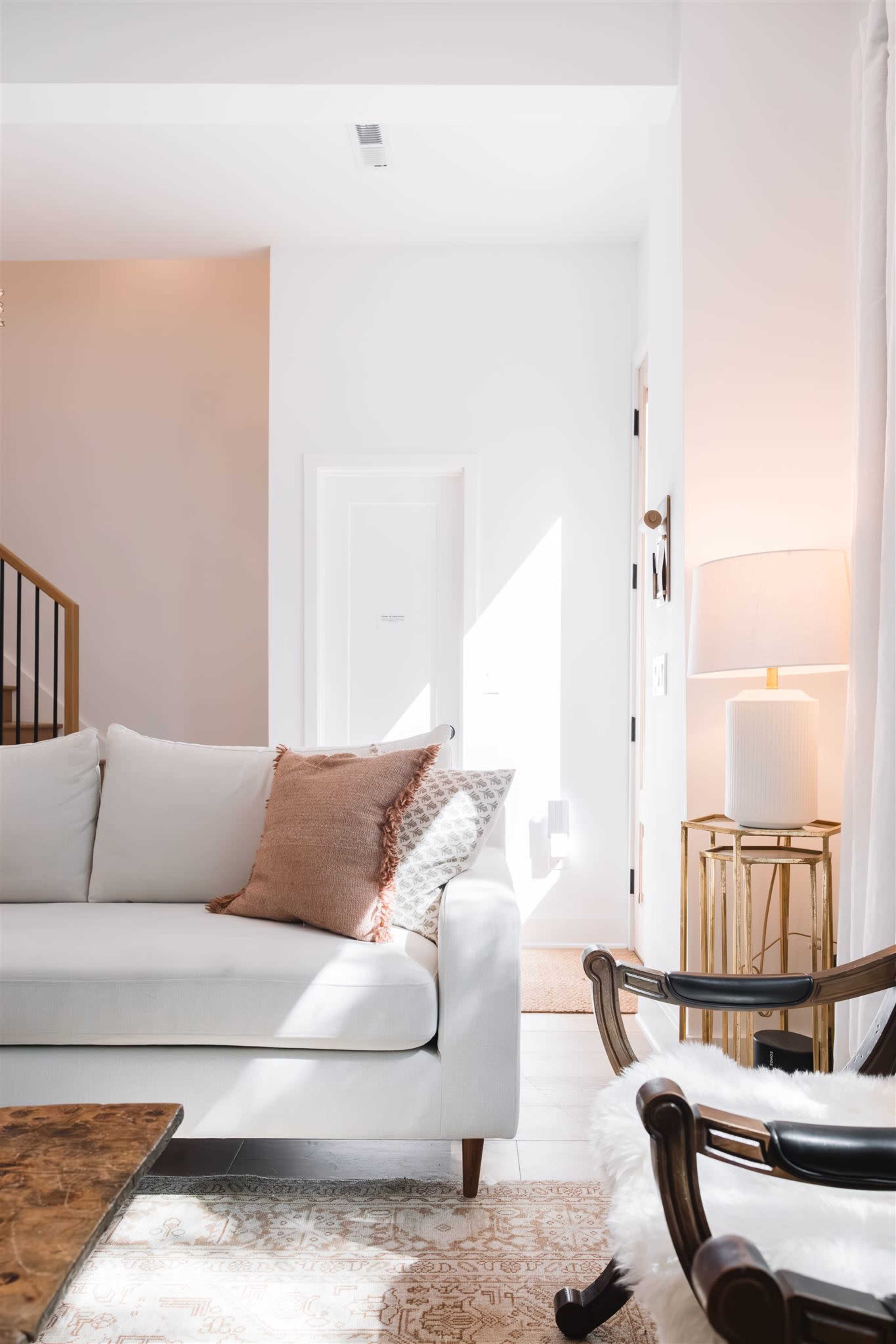 The image shows a bright living room with a white sofa adorned with a decorative pillow, a wooden coffee table, and a stylish lamp beside a chair.