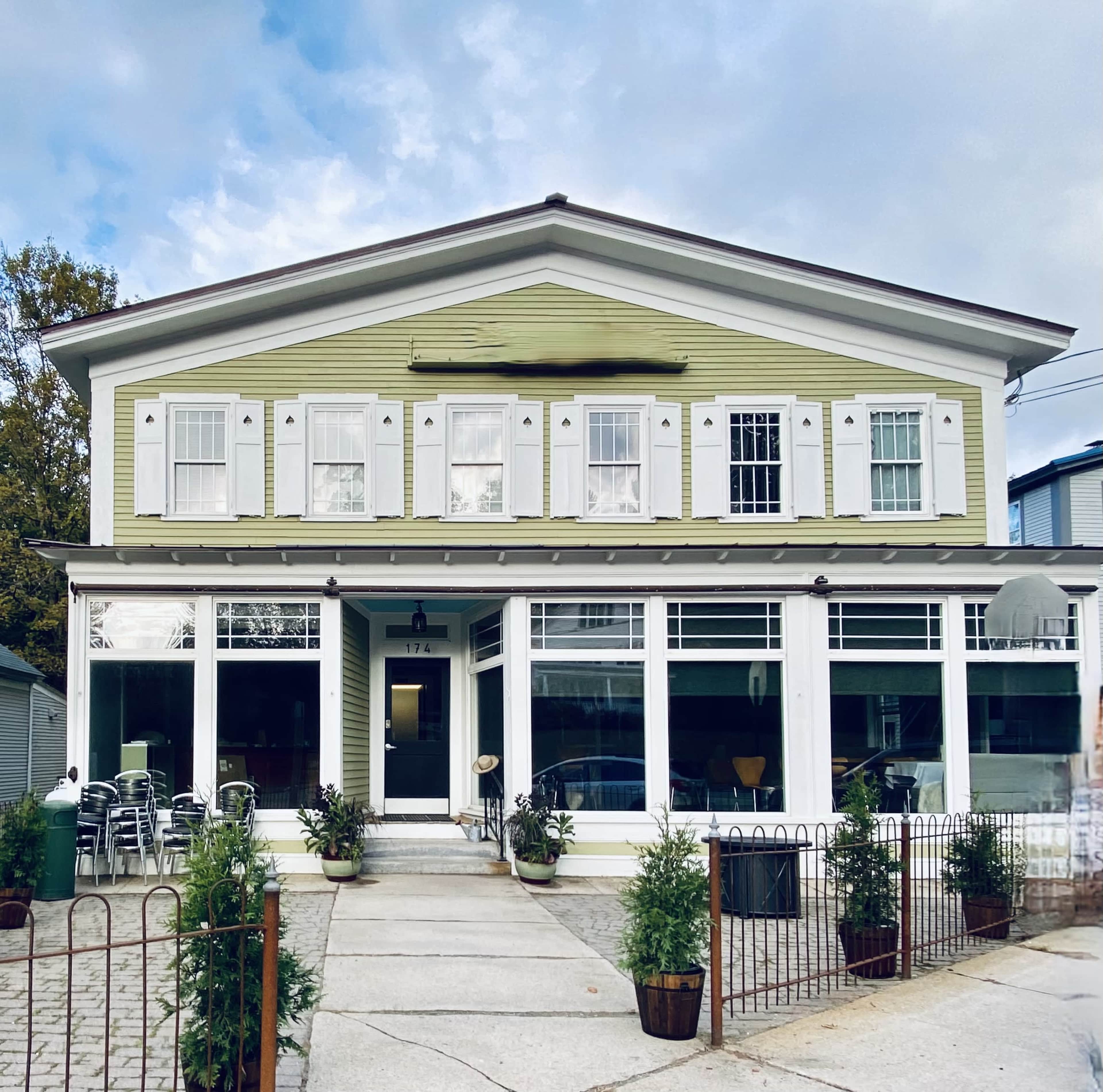 A large, two-story green house with white shutters and a covered porch, surrounded by a paved pathway and small planters.