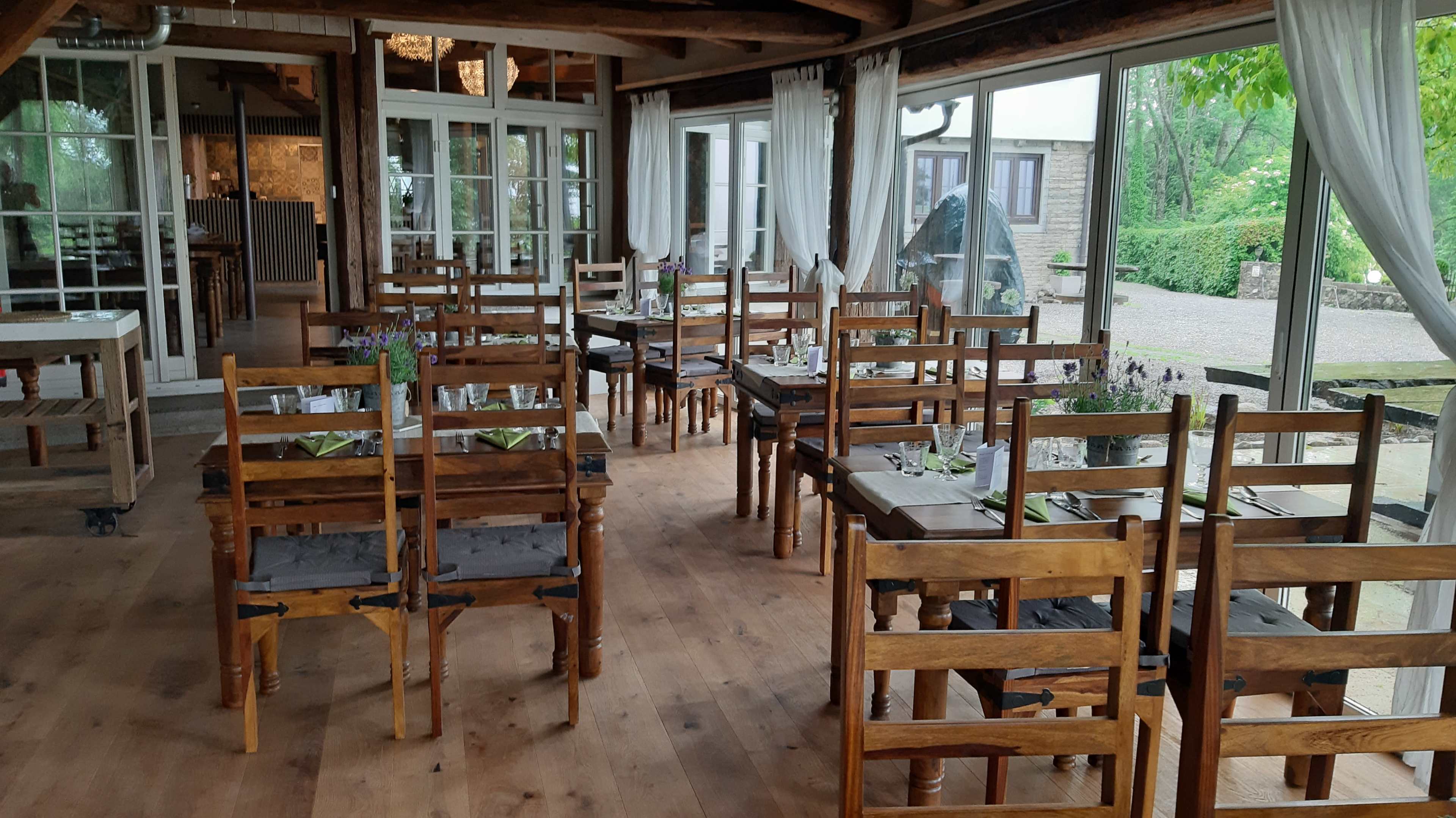 The image shows a wooden dining area with several tables and chairs arranged in a bright, glass-enclosed space surrounded by greenery.