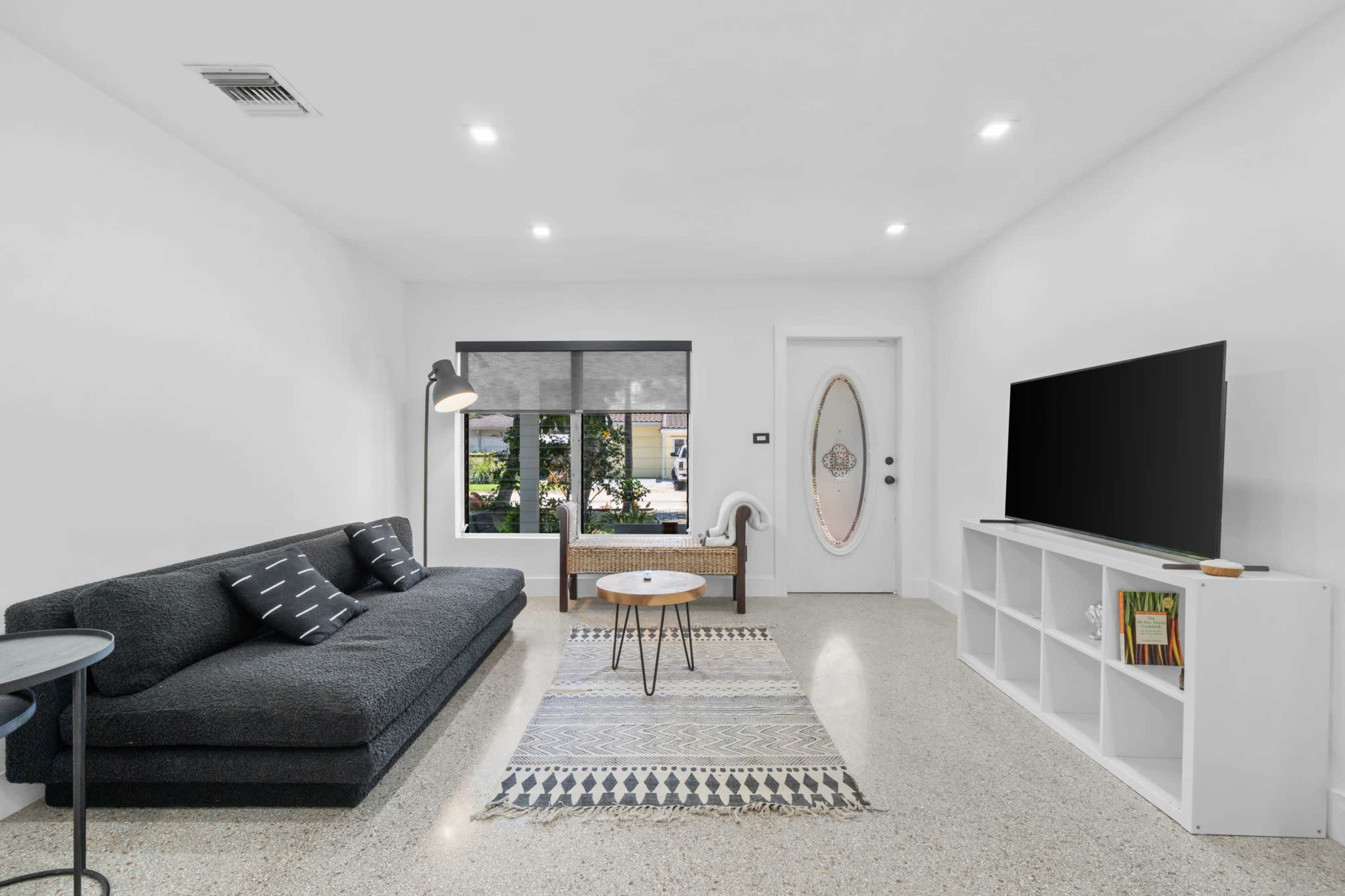 A modern living room features a dark sofa, a round coffee table, a television on a white media console, and a large window providing natural light.