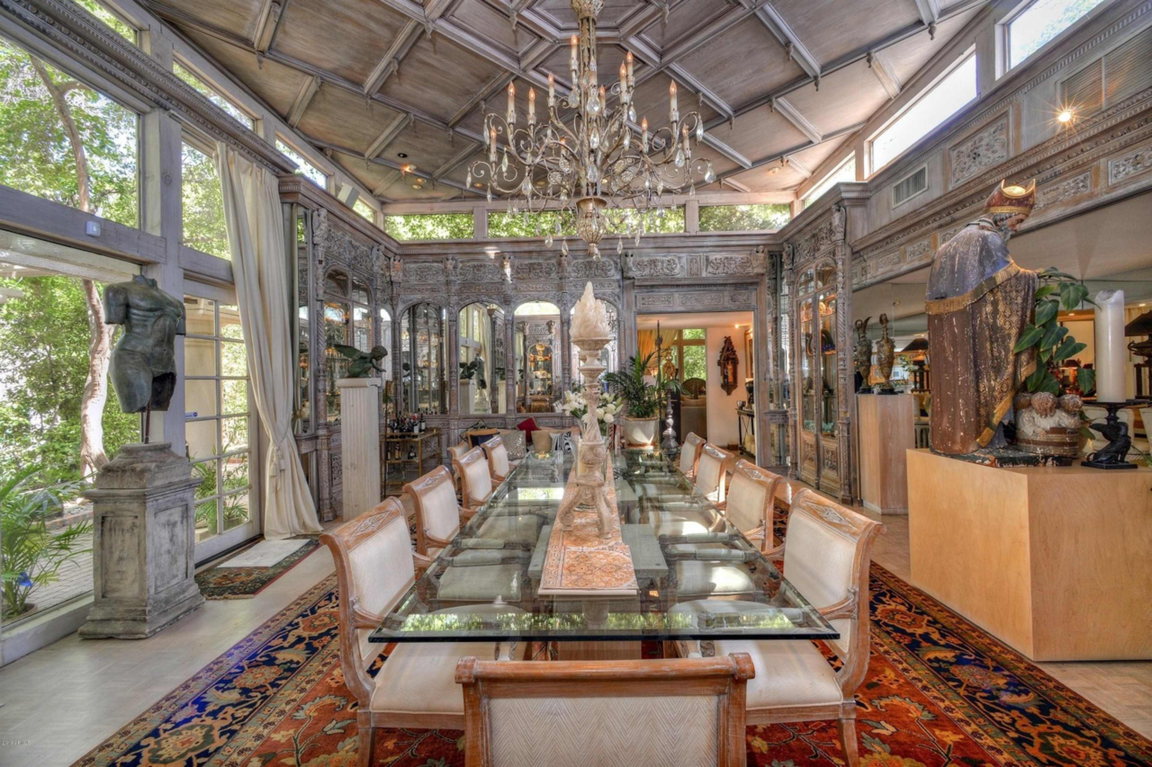 The image shows a spacious dining room with a long glass table, surrounded by chairs, and adorned with various sculptures and decorative elements.