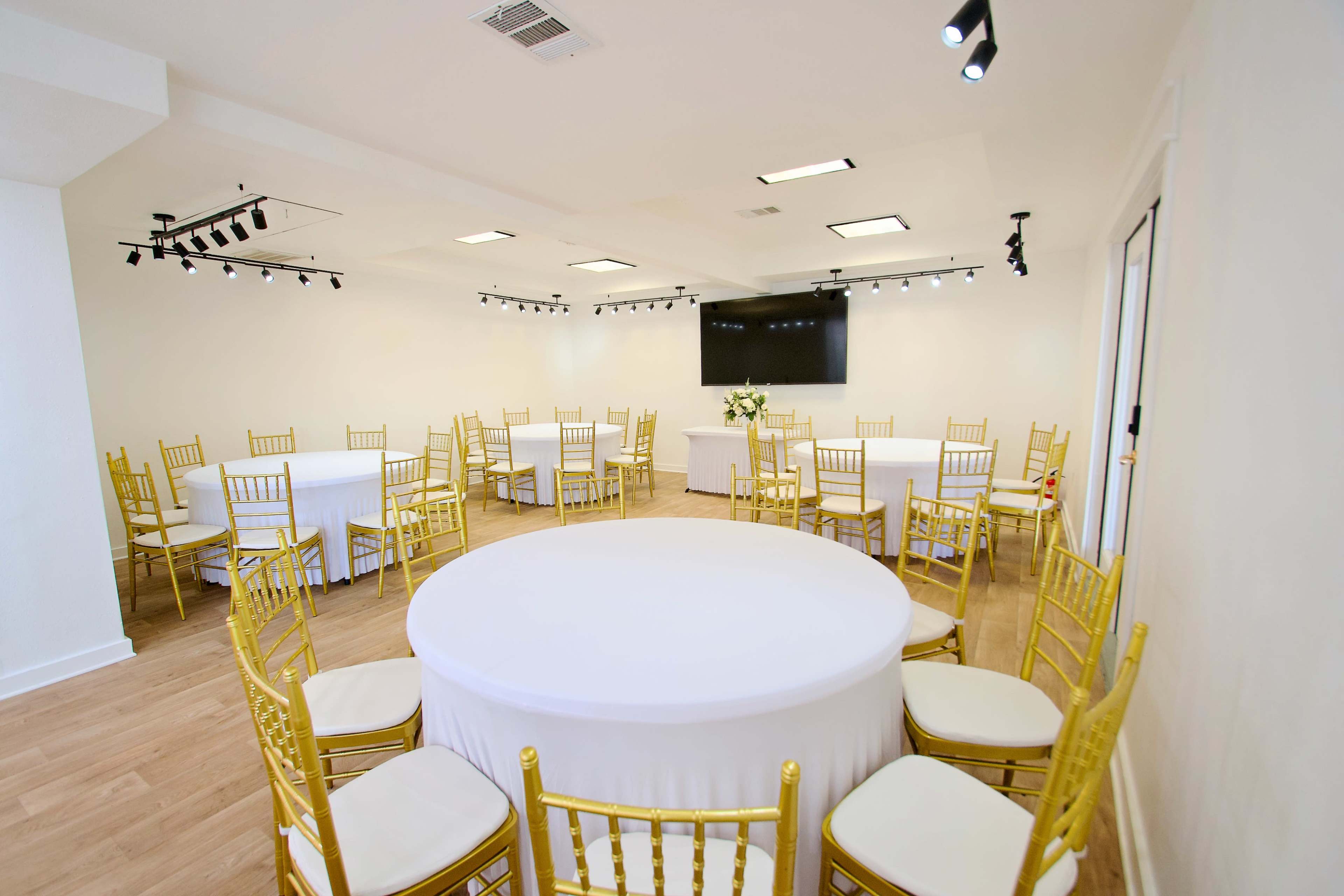 The image shows a well-lit event space featuring several round and rectangular tables covered with white tablecloths, accompanied by gold chairs arranged around them.