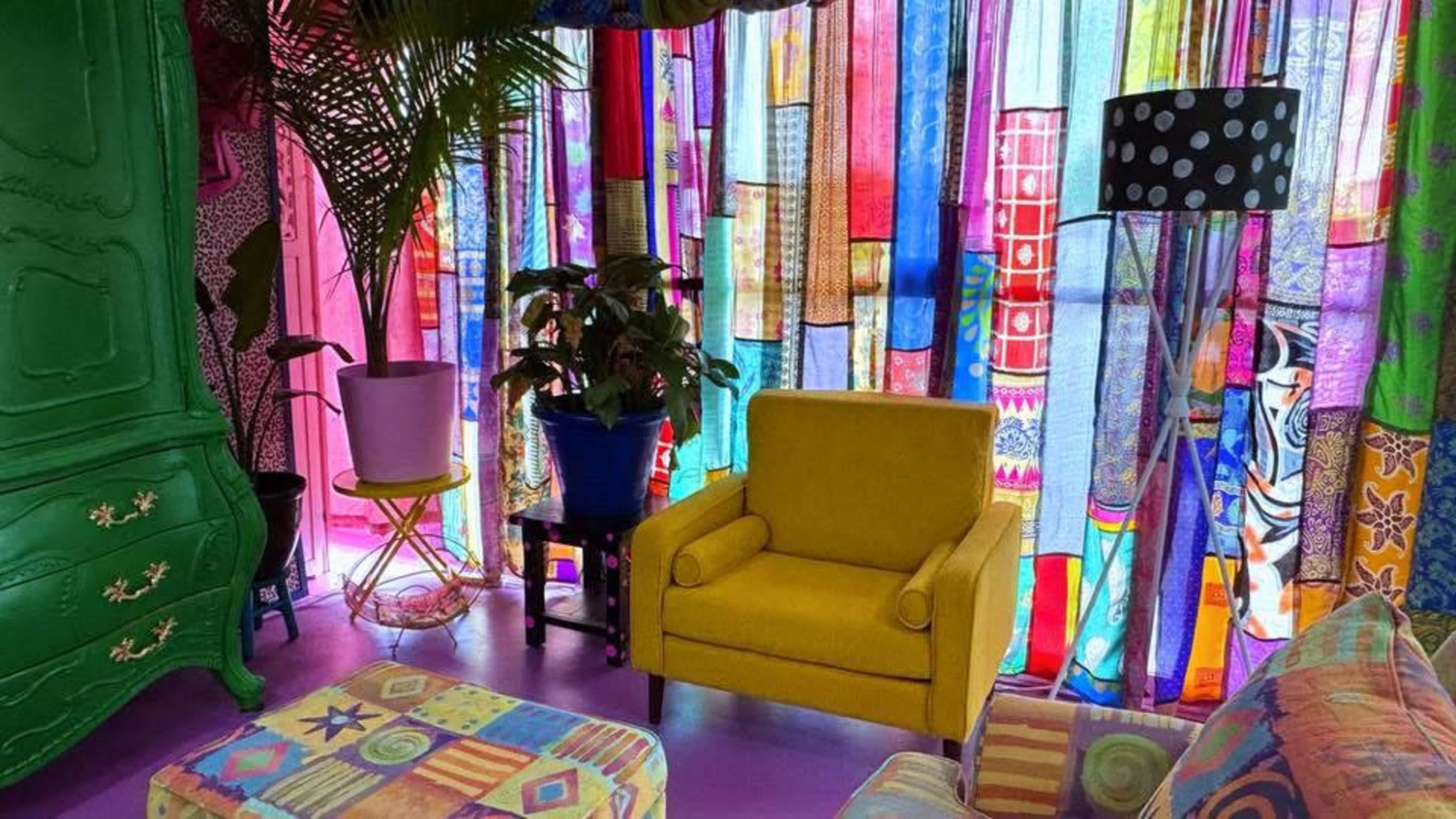 The image shows a vibrant room decorated with colorful fabric curtains, a yellow armchair, and a green cabinet, alongside potted plants and patterned furniture.