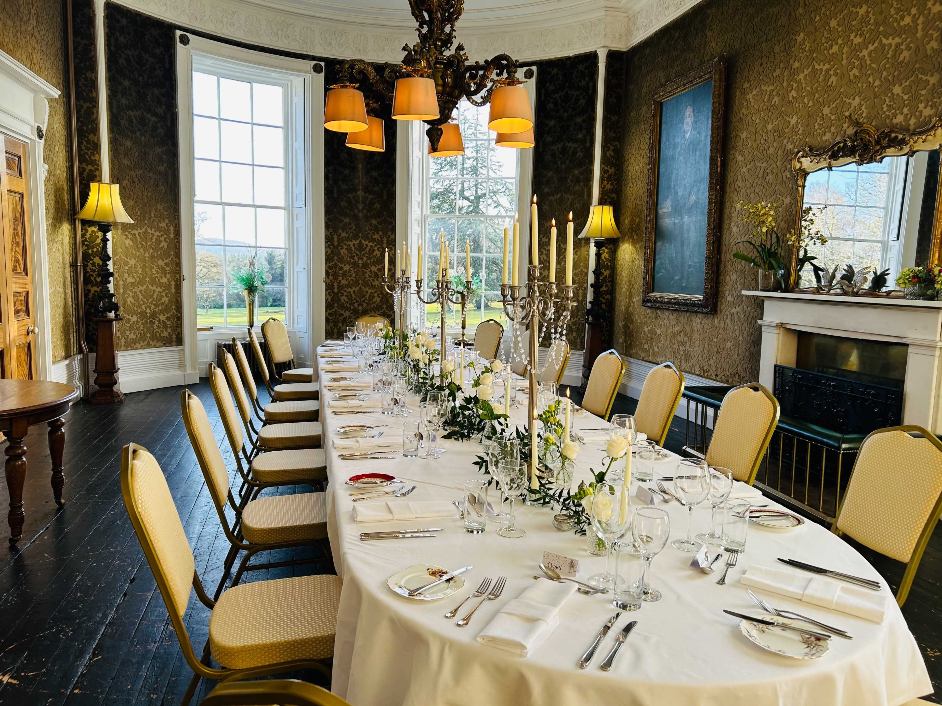 A long dining table is set with fine china, glassware, and candelabras in a elegantly decorated room with large windows and ornate wallpaper.