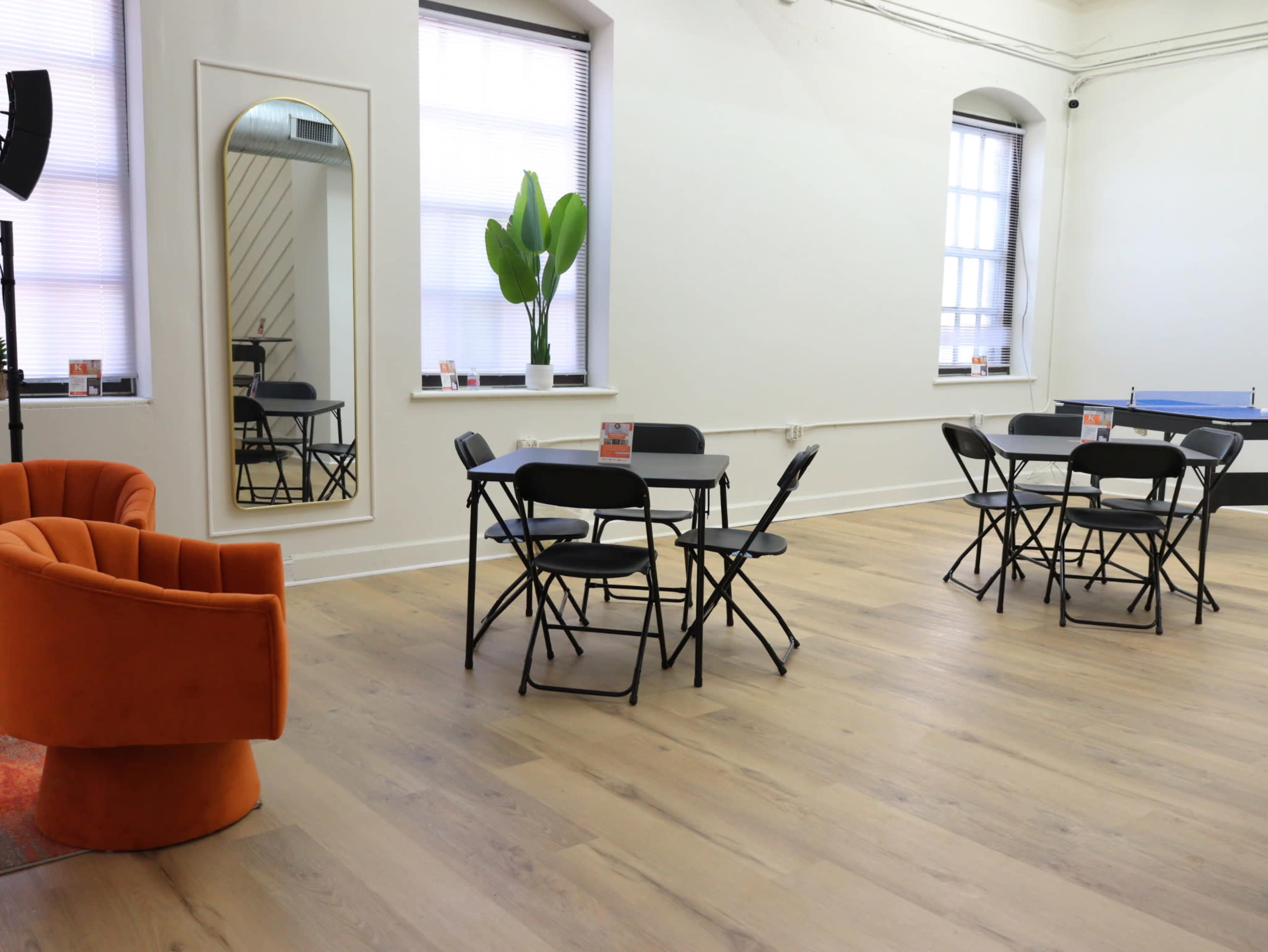 The image shows a brightly lit room with several black folding tables and chairs, a vibrant orange armchair, and a large potted plant near the window.