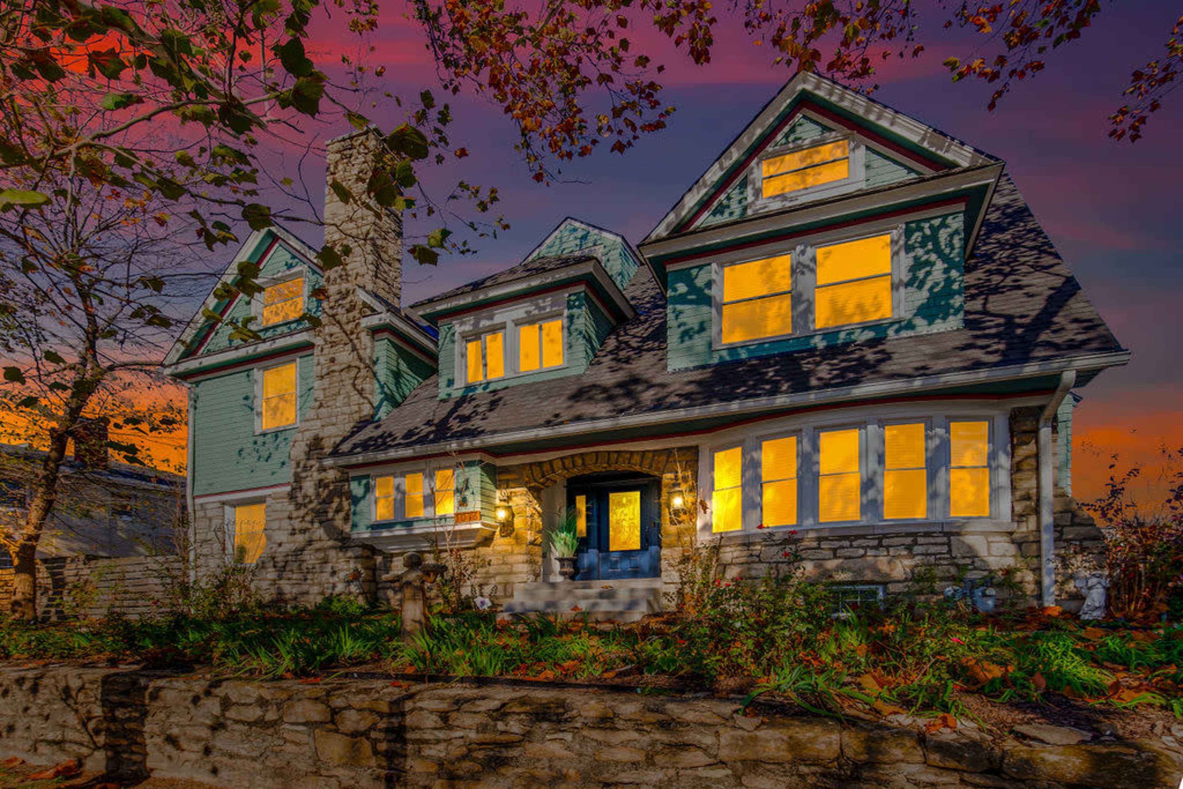 A large, two-story house with a stone facade and prominent windows is illuminated against a colorful sunset sky.