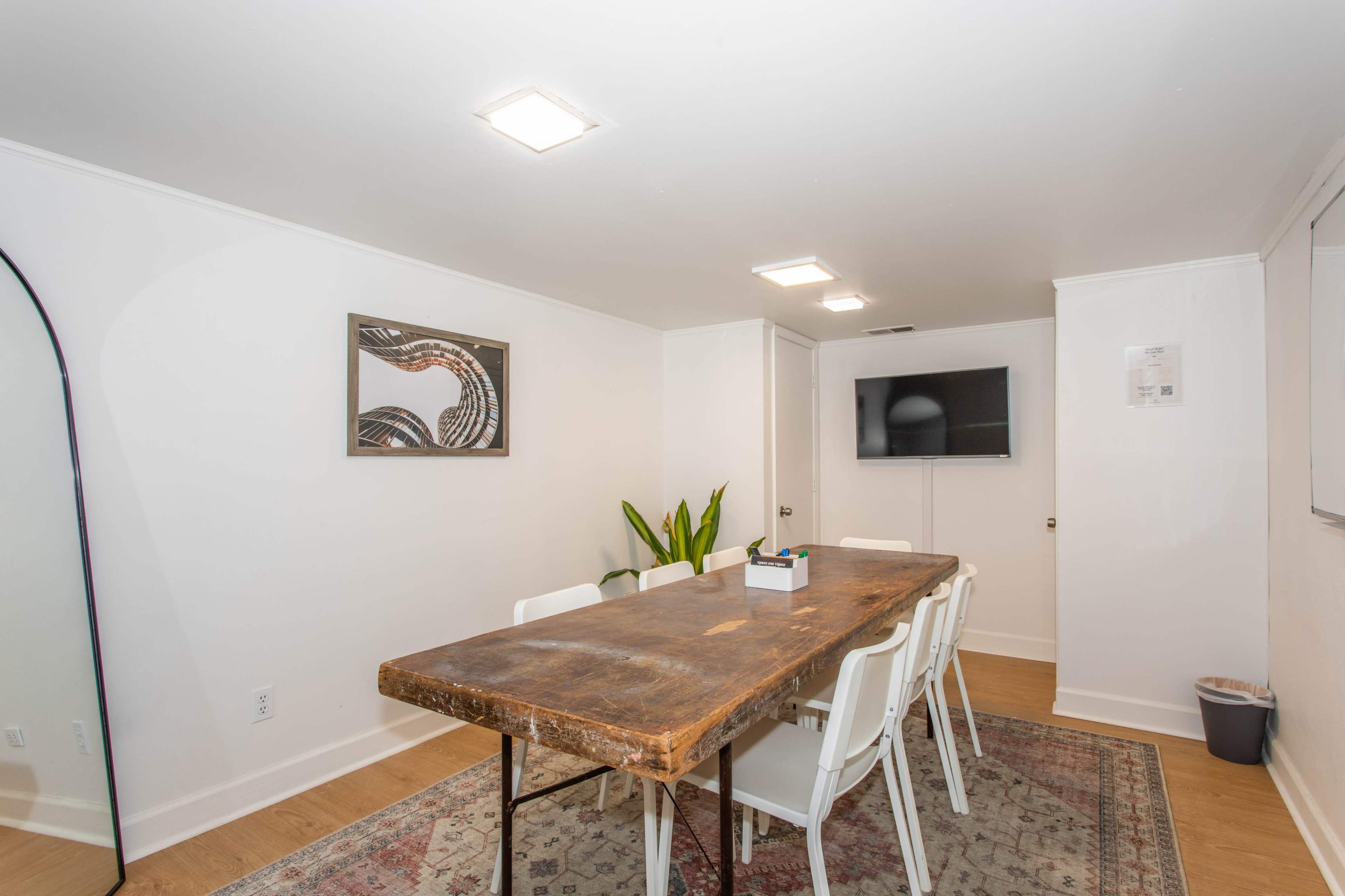A spacious room features a long wooden table surrounded by white chairs, with a wall-mounted television and a large mirror.