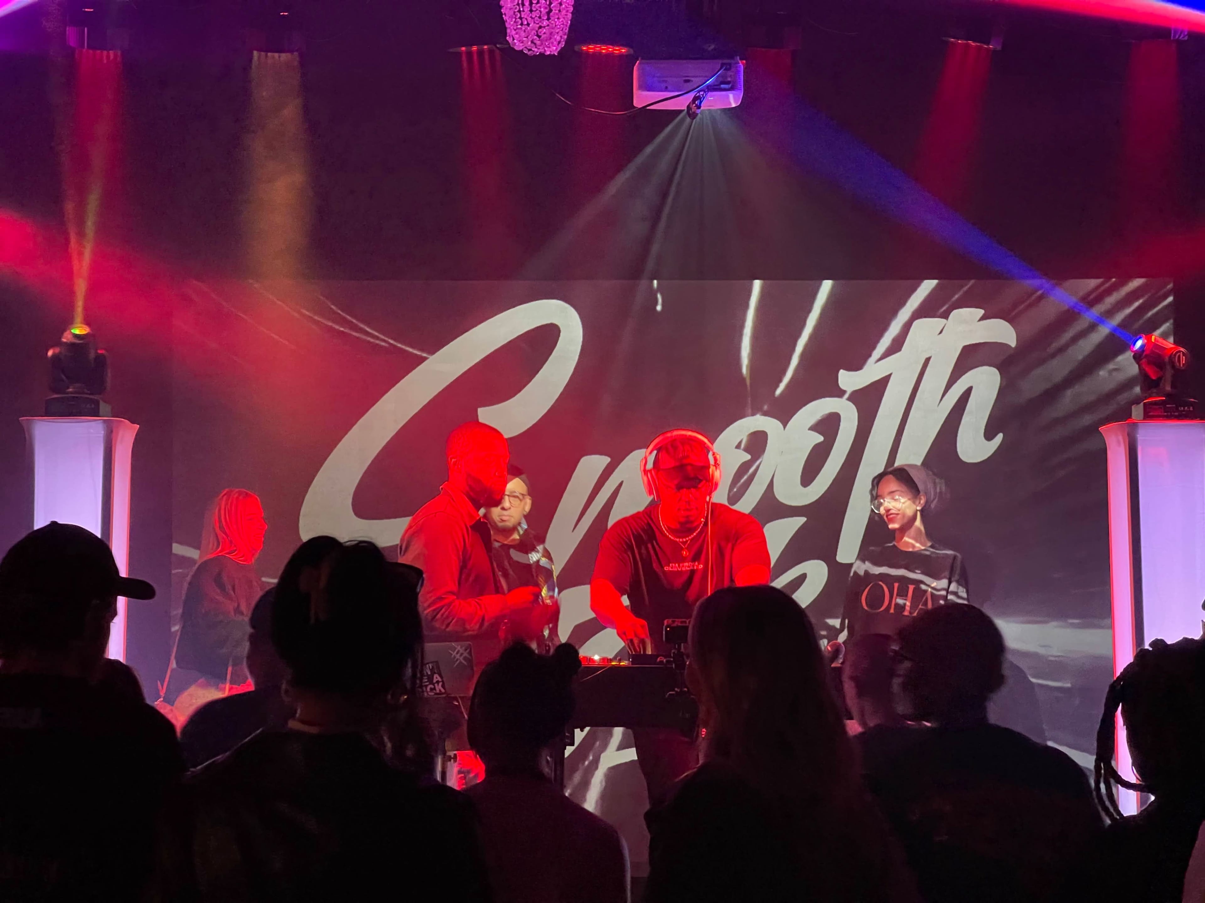A DJ performs on stage in front of a crowd with colorful lighting and a backdrop displaying the word "Smooth."