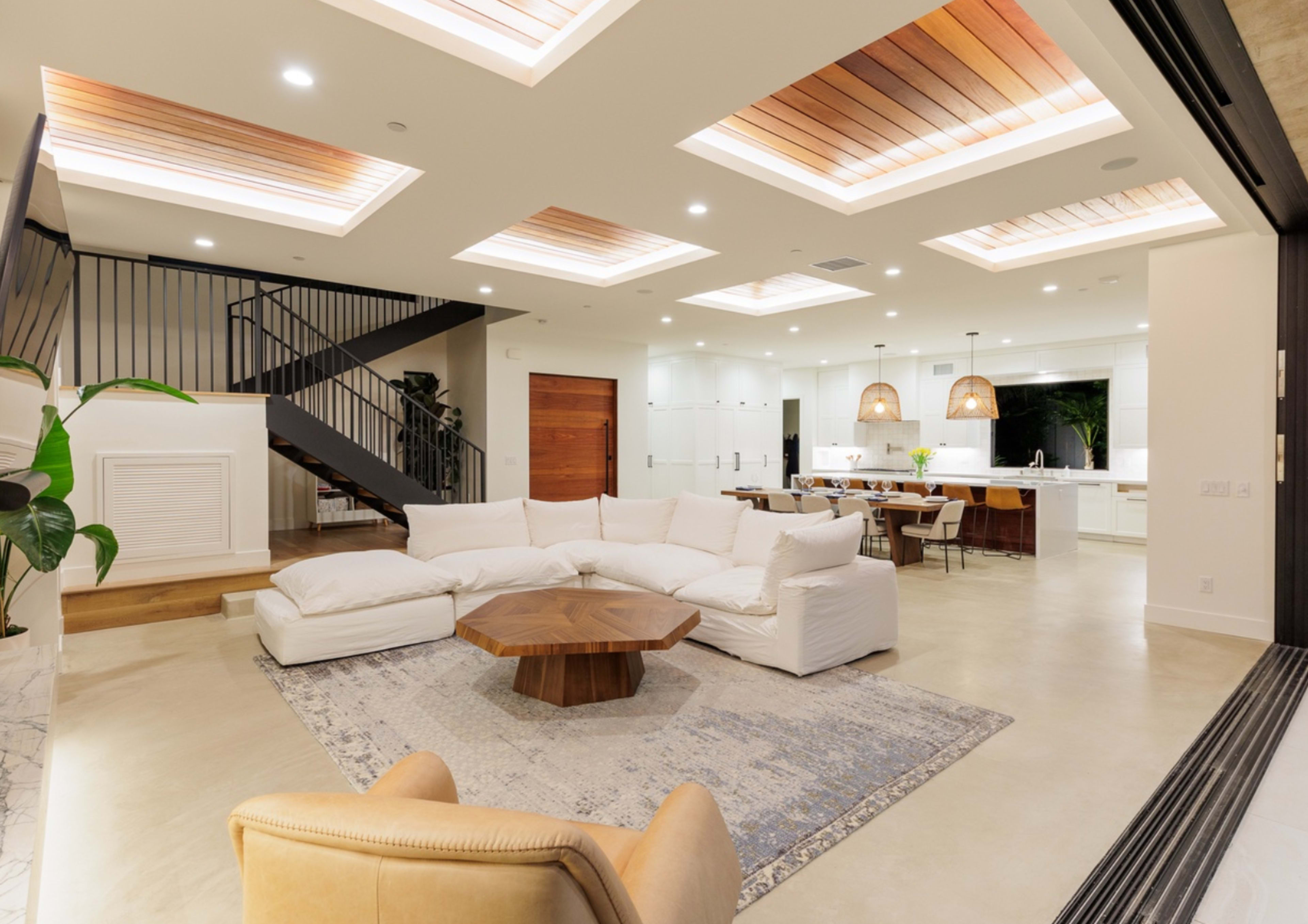 A modern living room with a white sectional sofa, a wooden coffee table, and a staircase leading to an upper level, adjacent to a dining area and kitchen.