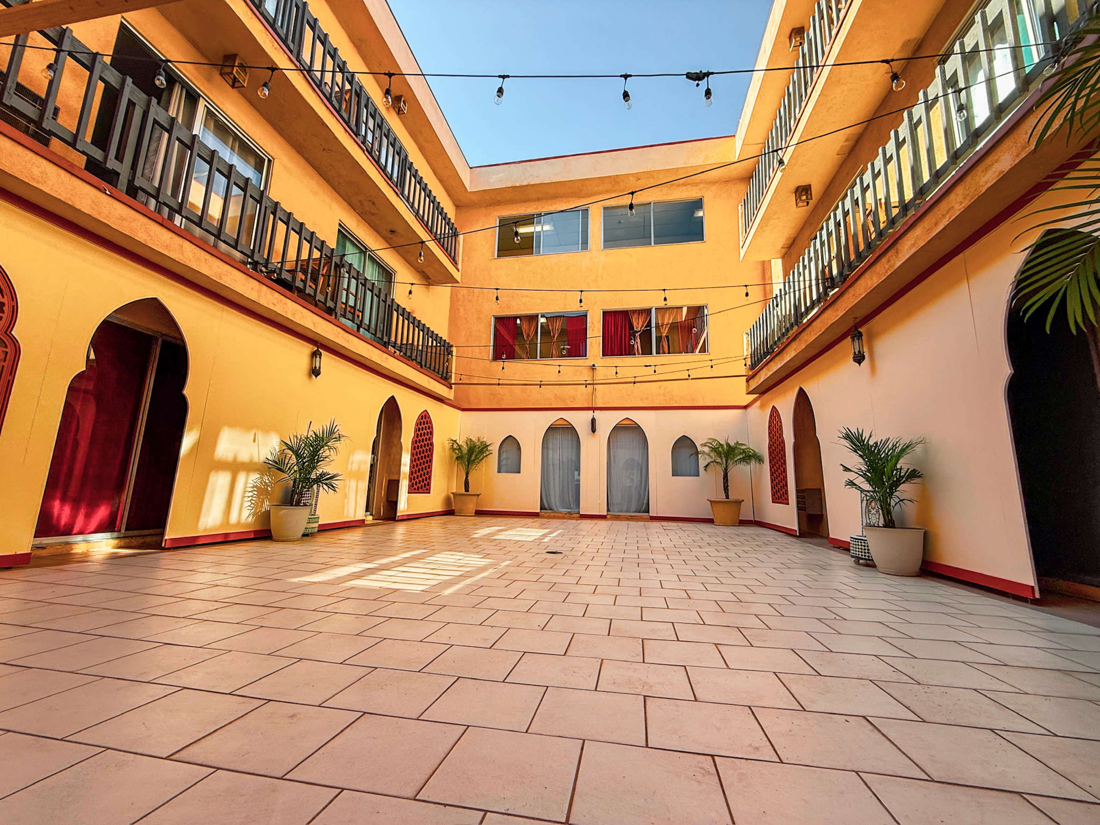 The image shows a spacious, open courtyard surrounded by buildings with balconies, featuring decorative arches and potted plants.