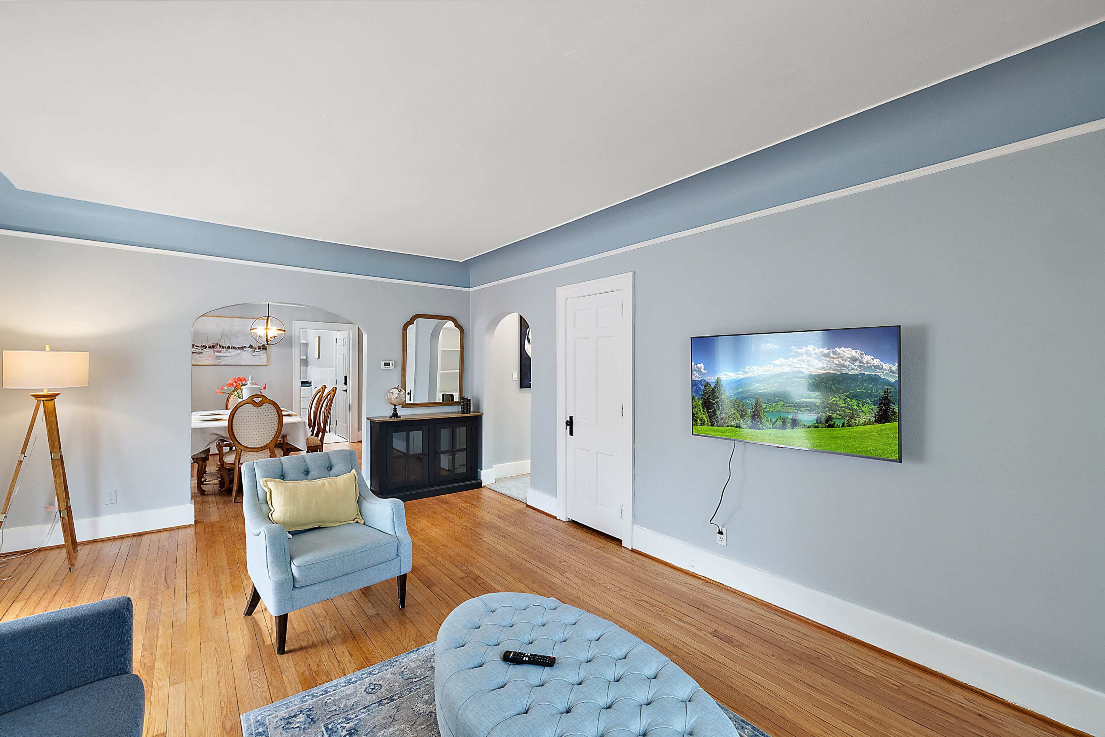 A living room with a light blue wall, a mounted television displaying a landscape, a light blue armchair, an ottoman, and a view into a dining area with a table and chairs.