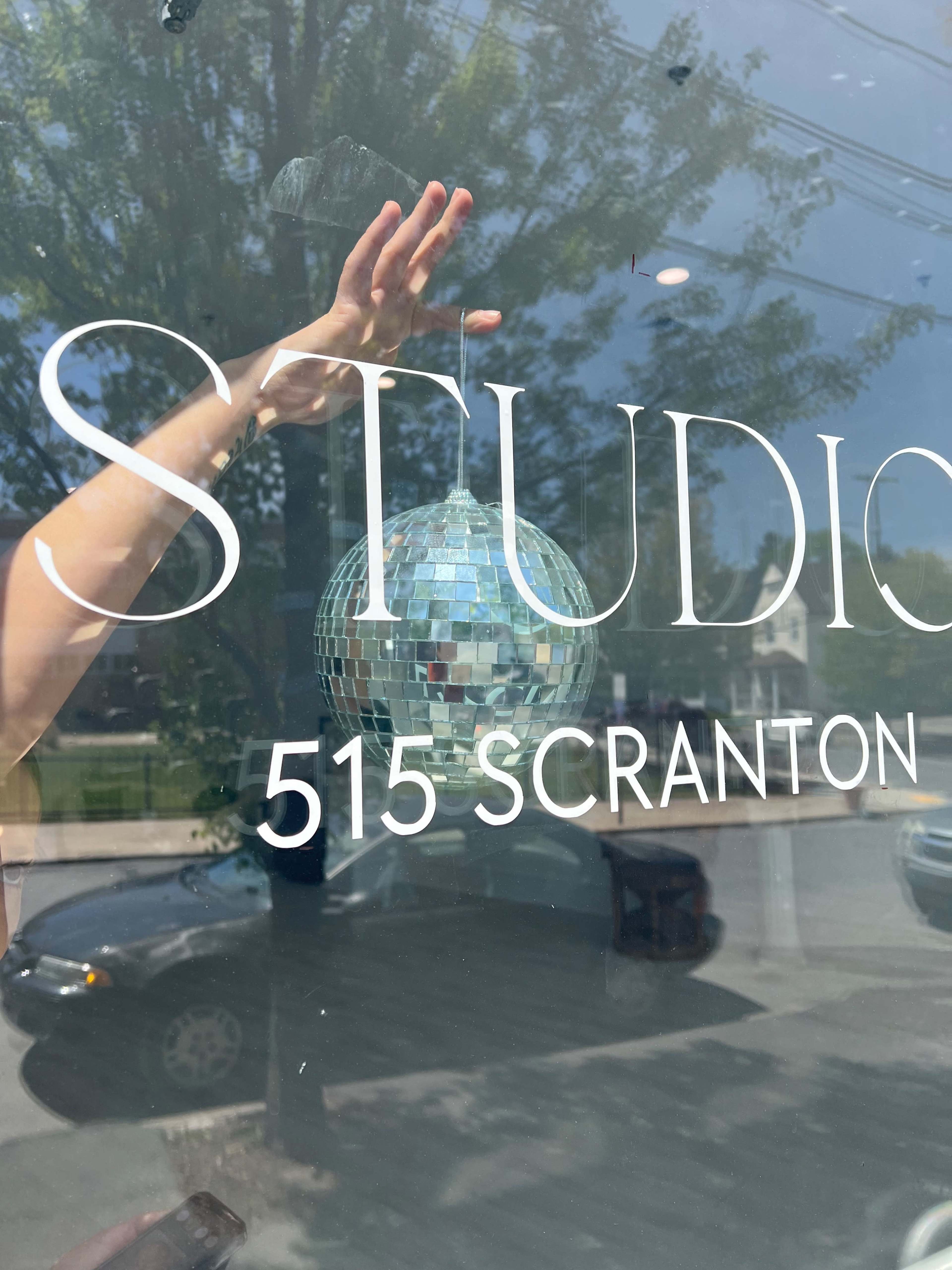 A hand reaches out to touch a disco ball hanging in front of a storefront window displaying the name "STUDIO" and the address "515 SCRANTON."