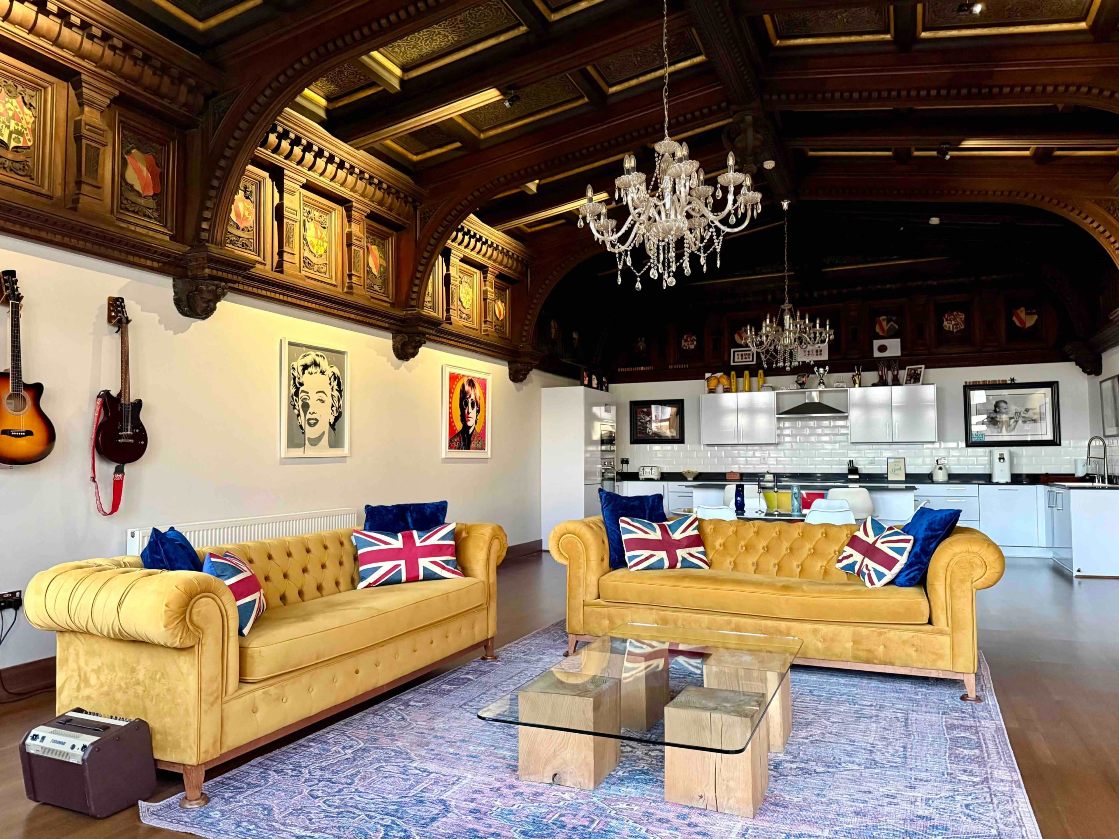 A spacious living area features two yellow chesterfield sofas adorned with Union Jack pillows, a glass coffee table, and a kitchen area in the background with modern appliances.