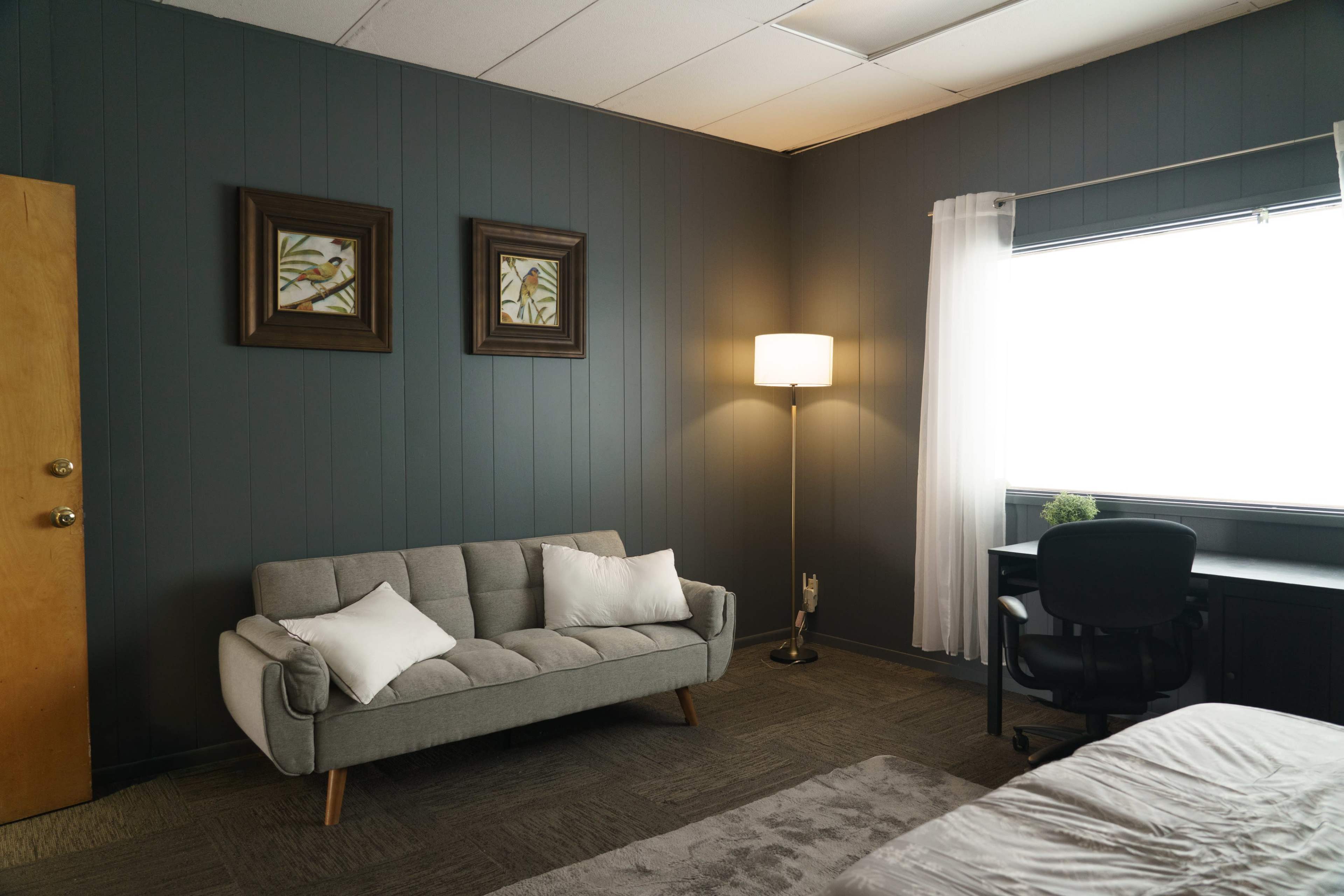 The room features a gray couch with white pillows, a floor lamp, a desk with a chair, and artwork on the walls.