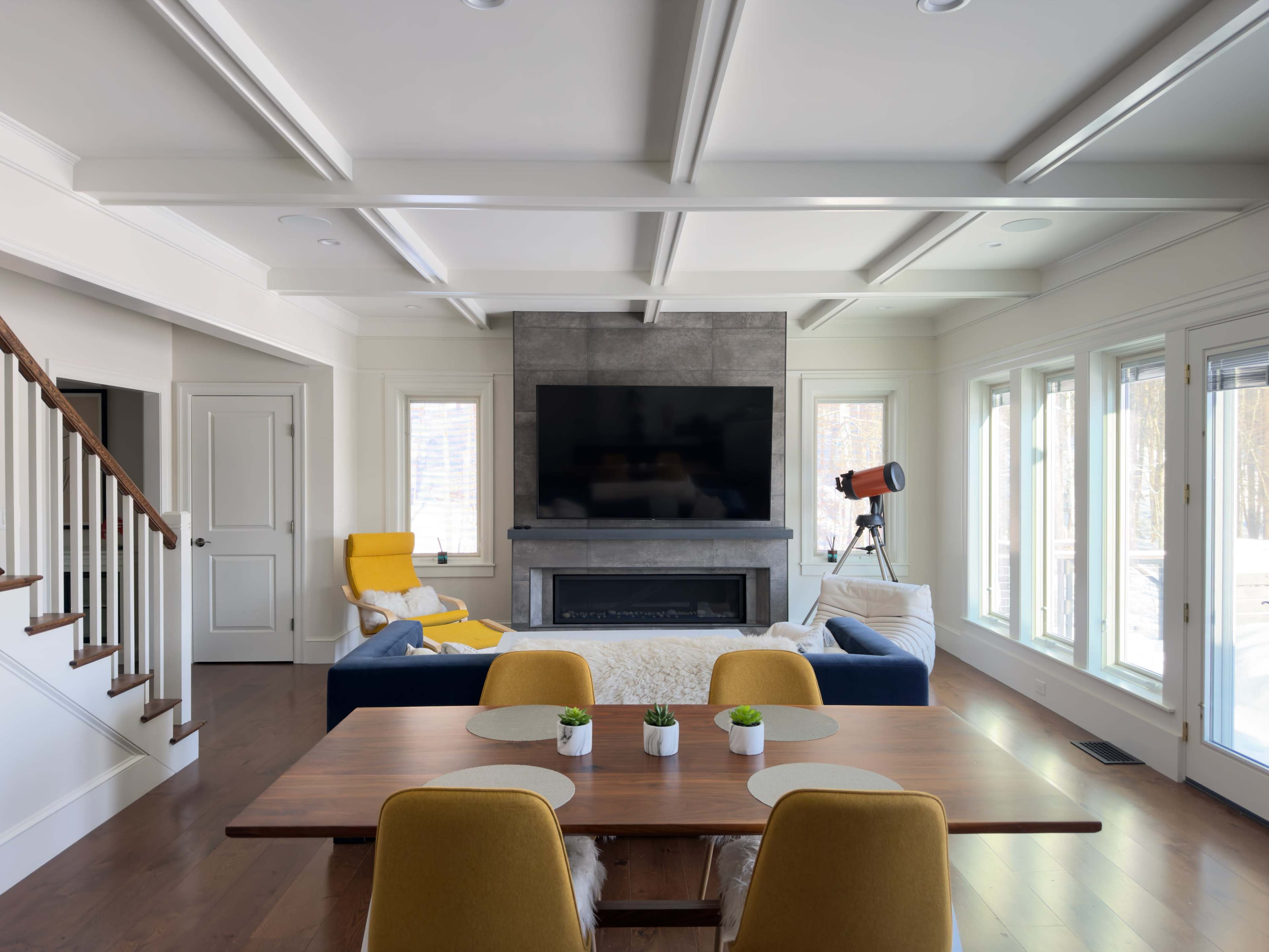 The image shows a modern living room with a gray couch, a large television mounted on the wall, and a dining area featuring a wooden table and yellow chairs.