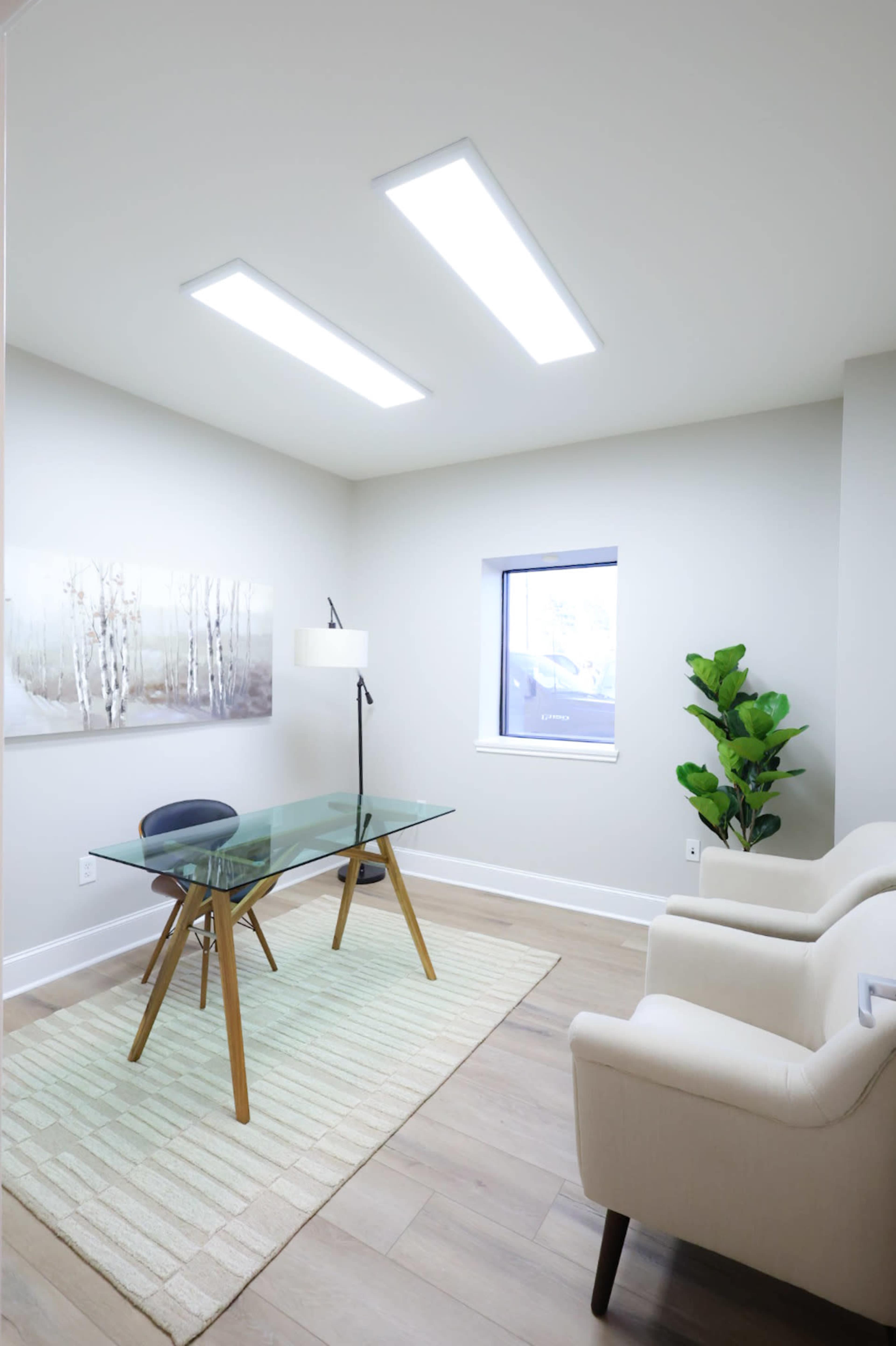 A modern office space features a glass desk with wooden legs, a white armchair, a floor lamp, and a small plant, complemented by artwork on the wall and a window letting in natural light.