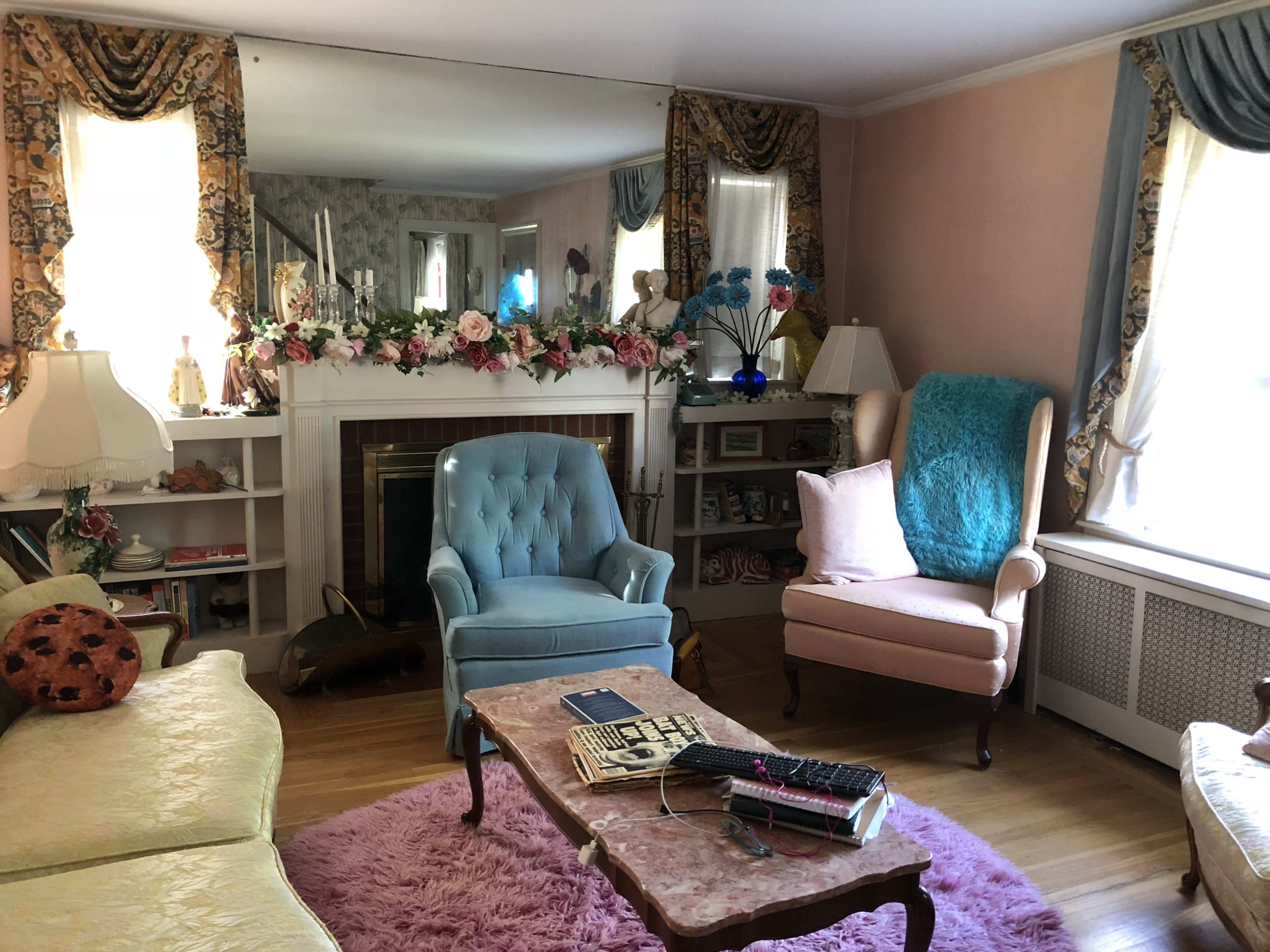 The room features a pastel color scheme with a pink rug, two upholstered chairs, a floral arrangement on the mantel, and a large mirror reflecting the space.
