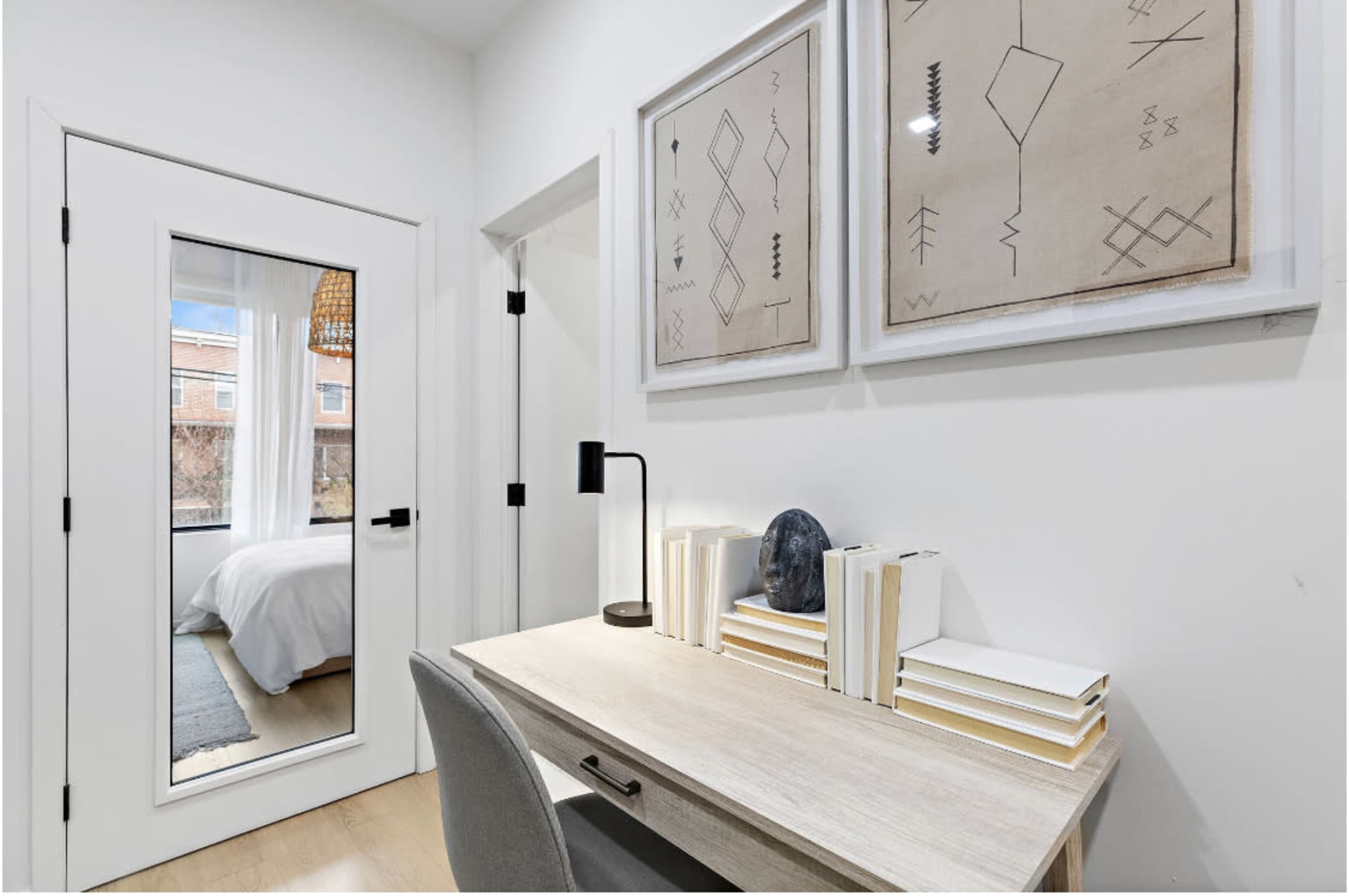 A small, minimalist office space with a desk, stack of books, a lamp, and two framed artworks on the wall, adjacent to a door leading to another room.