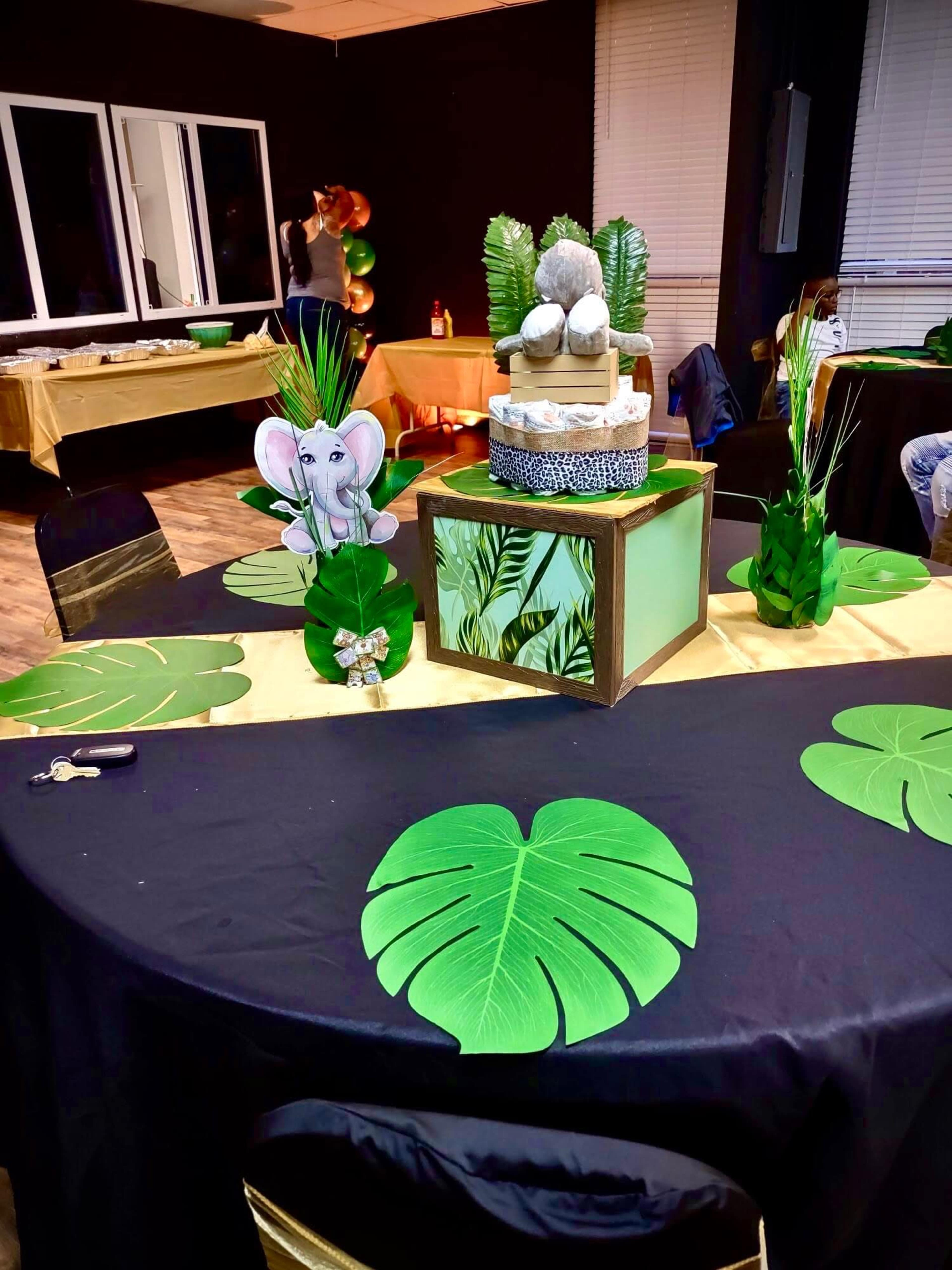 A decorated event space features a table set with green leaf-shaped decor, an elephant centerpiece, and a tiered cake surrounded by tropical foliage.