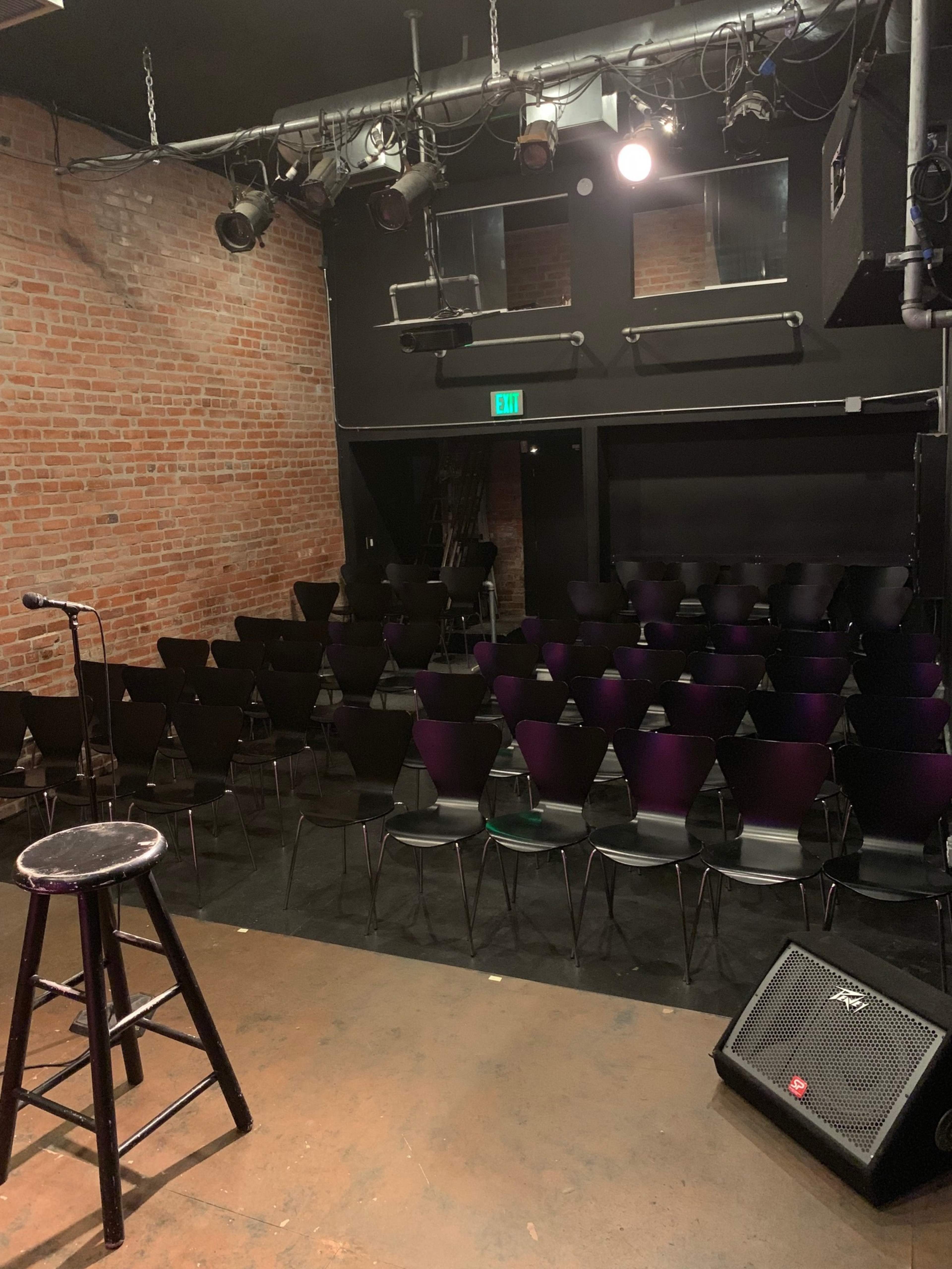 A small performance stage with a microphone and stool in front of a dark room filled with rows of black chairs.