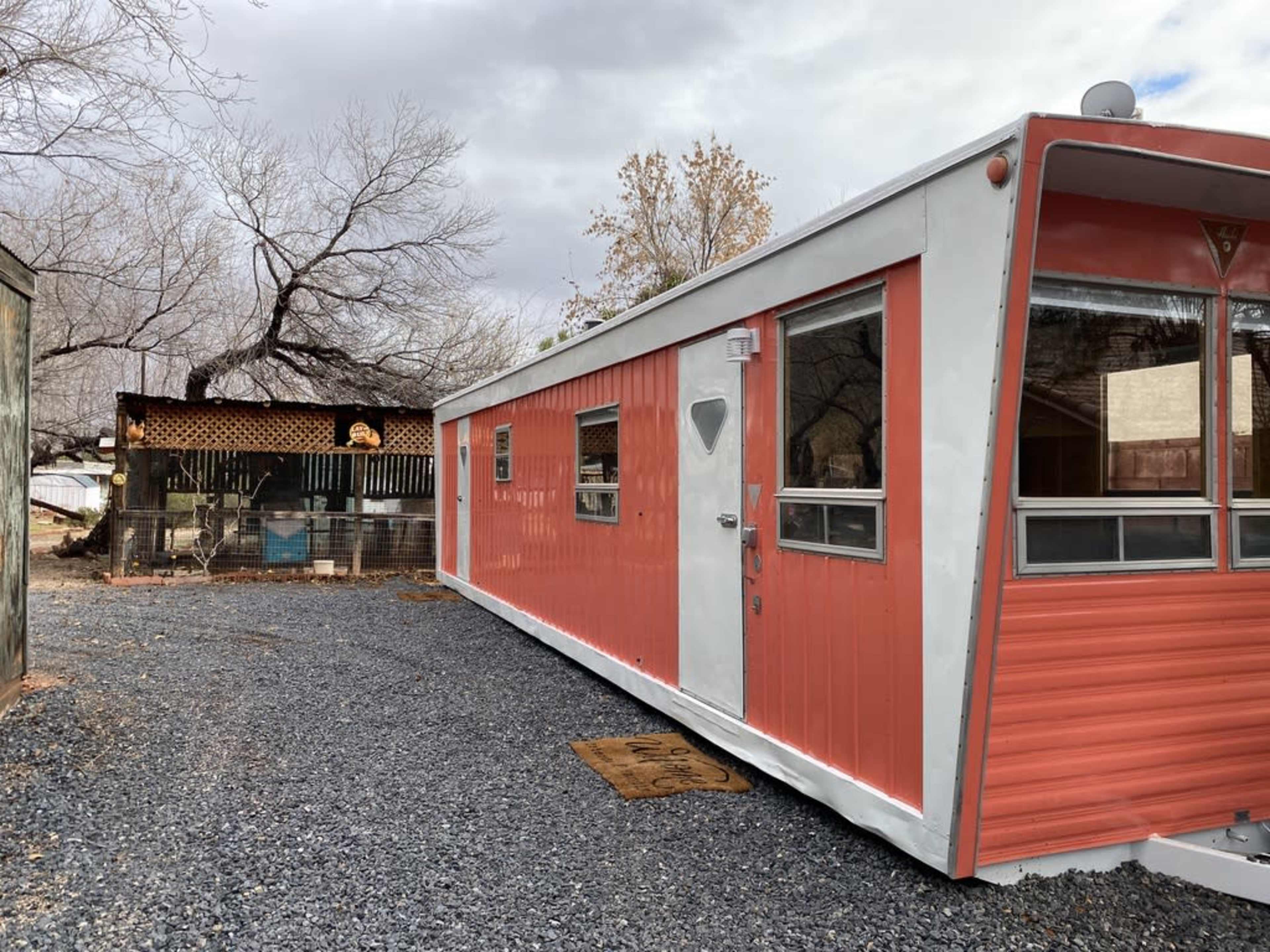 A retro-style mobile home with orange and white siding is parked on a gravel driveway next to a rustic structure in a treed outdoor setting.