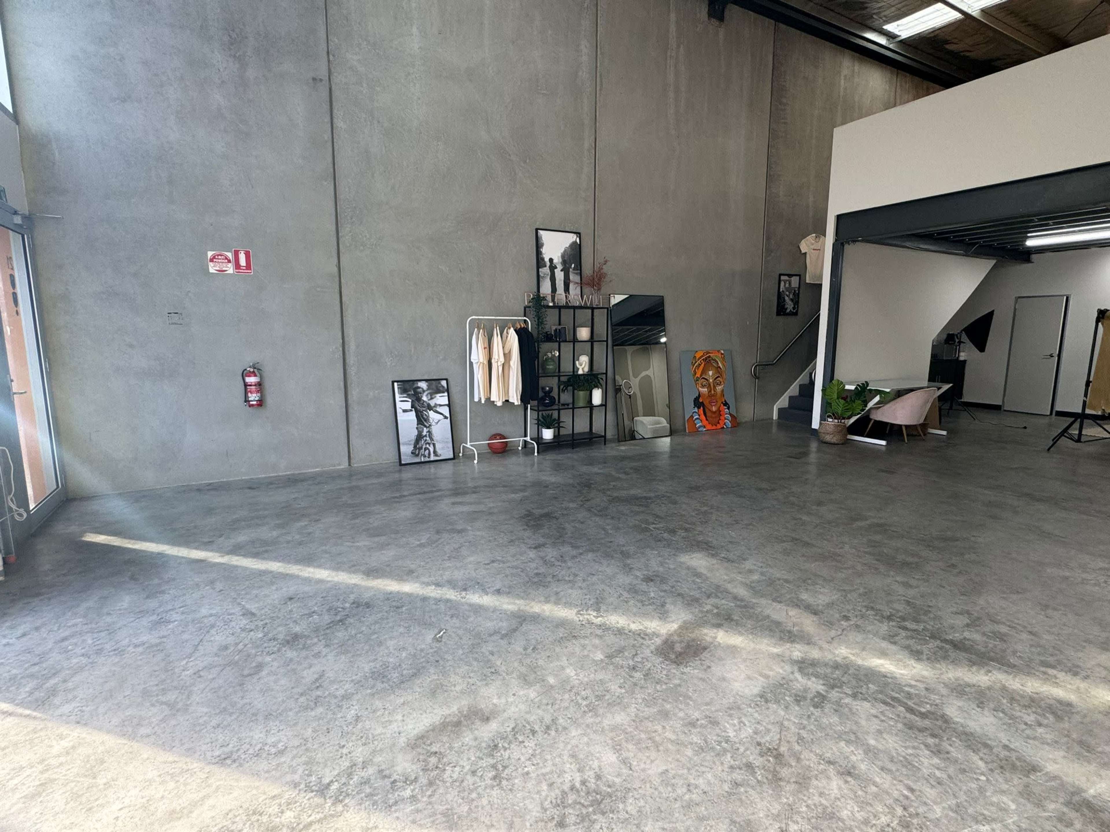 The interior of a spacious studio features a concrete floor, an industrial-style wall, various clothing displays, and an art piece against one wall.
