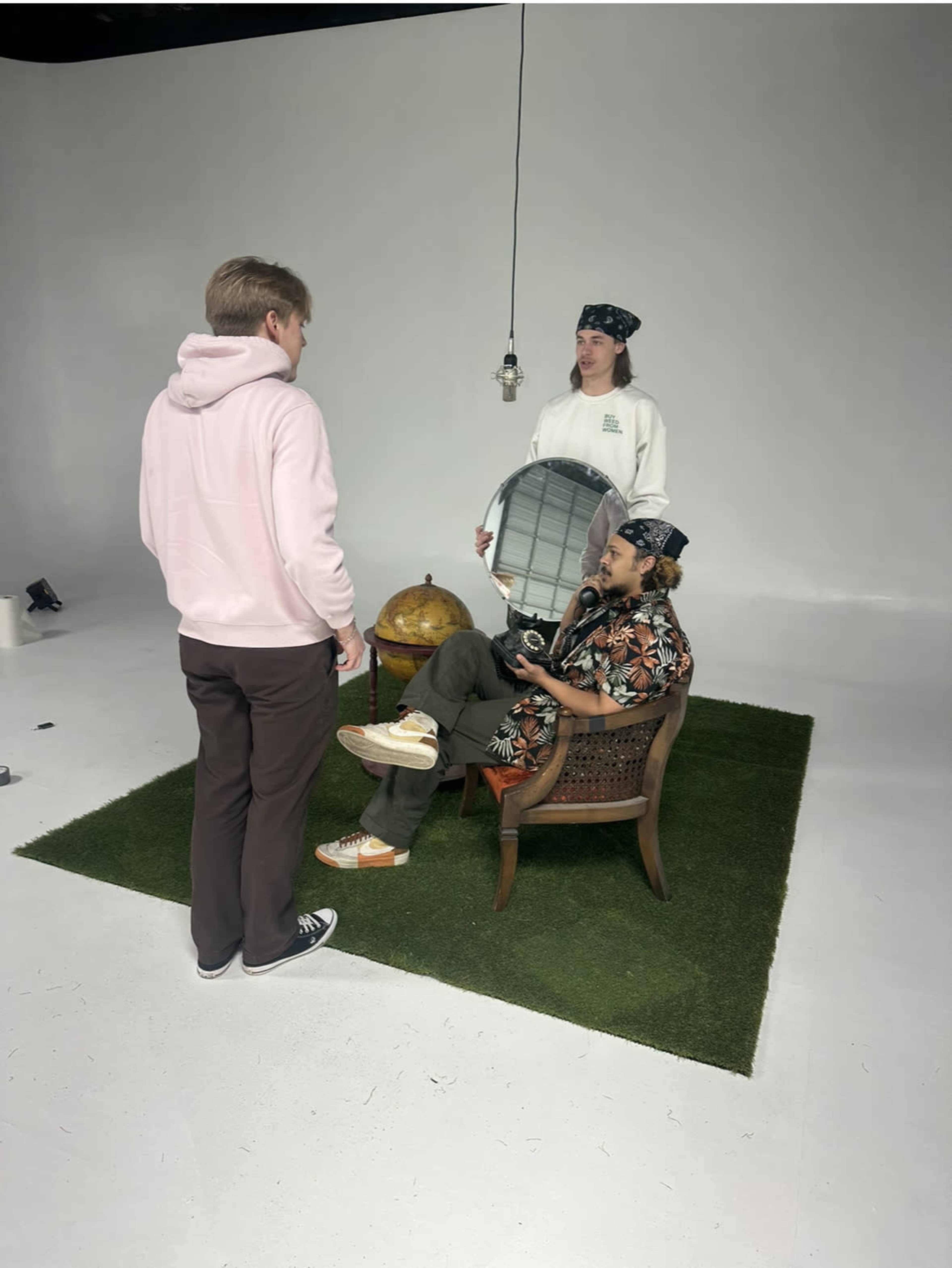 A group of three people are engaged in conversation in a studio setting, with one person seated on a chair and holding a camera, while another stands nearby holding a mirror.
