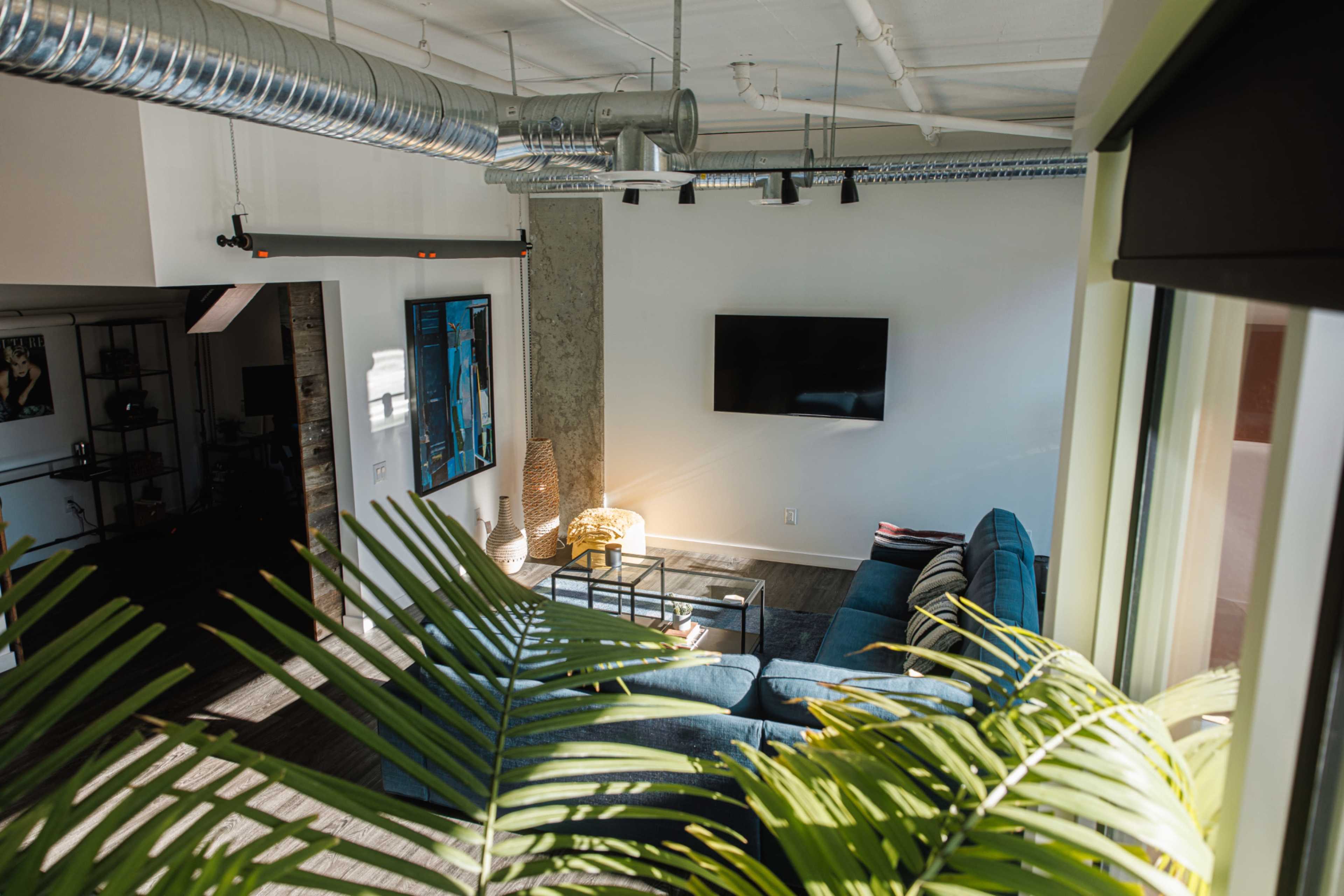 A modern living space features a blue sectional sofa, a wall-mounted TV, and large windows adorned with greenery.