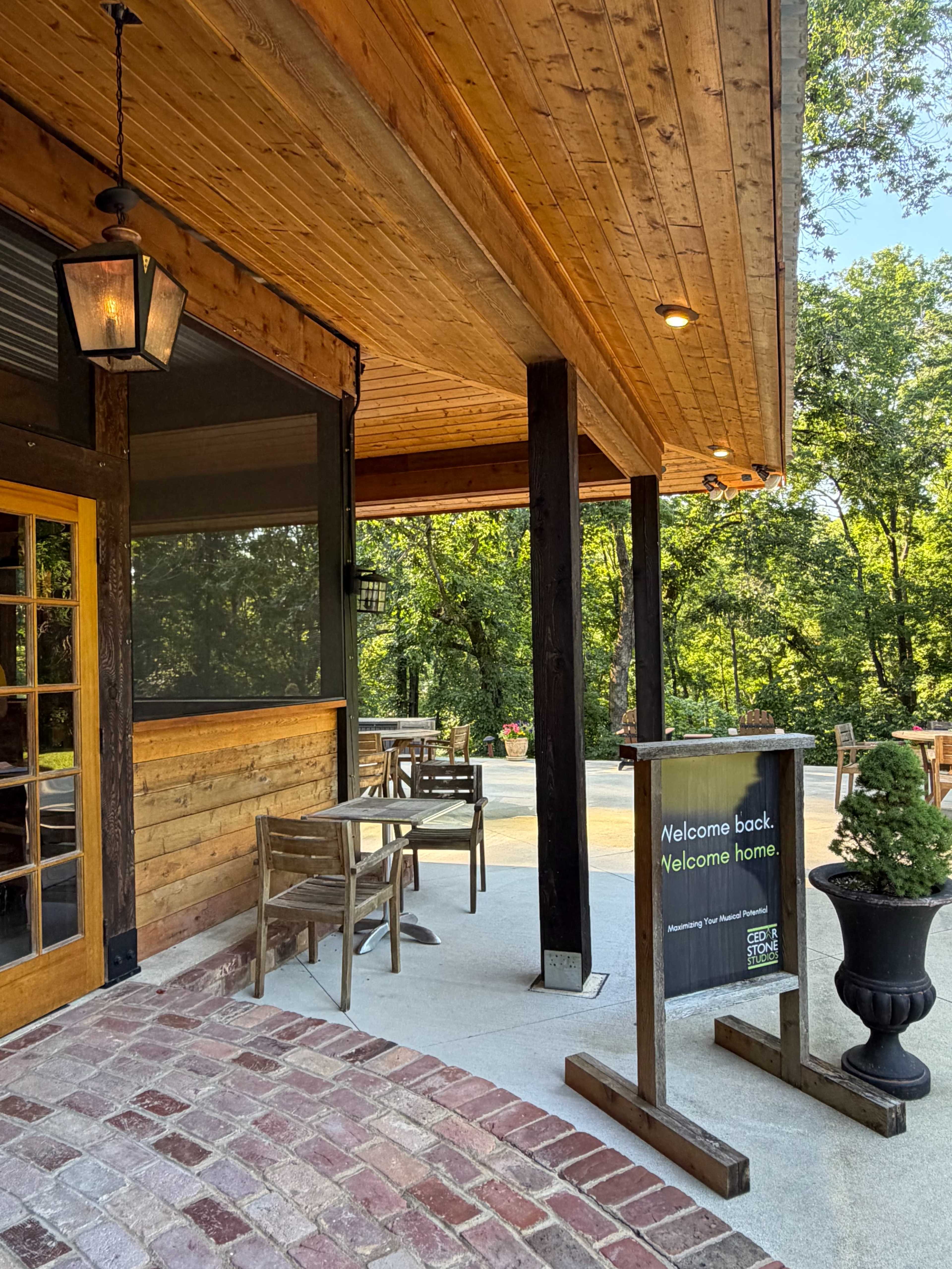 A wooden patio with tables and chairs is set outside a restaurant, featuring a welcoming sign and surrounded by trees.