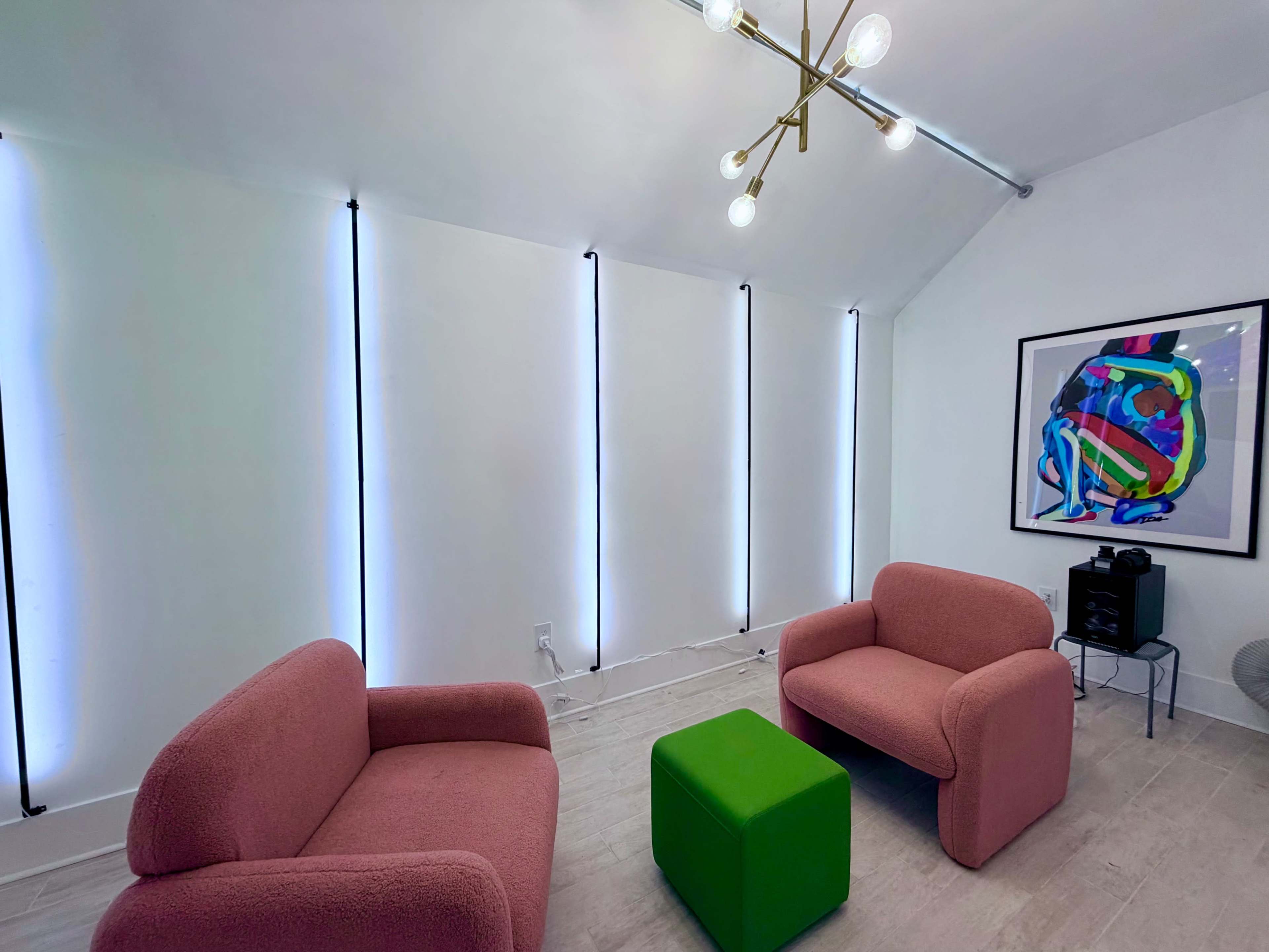 The room features two pink armchairs and a green cube-shaped table, with bright vertical lights illuminating the white walls and a colorful artwork displayed on one side.