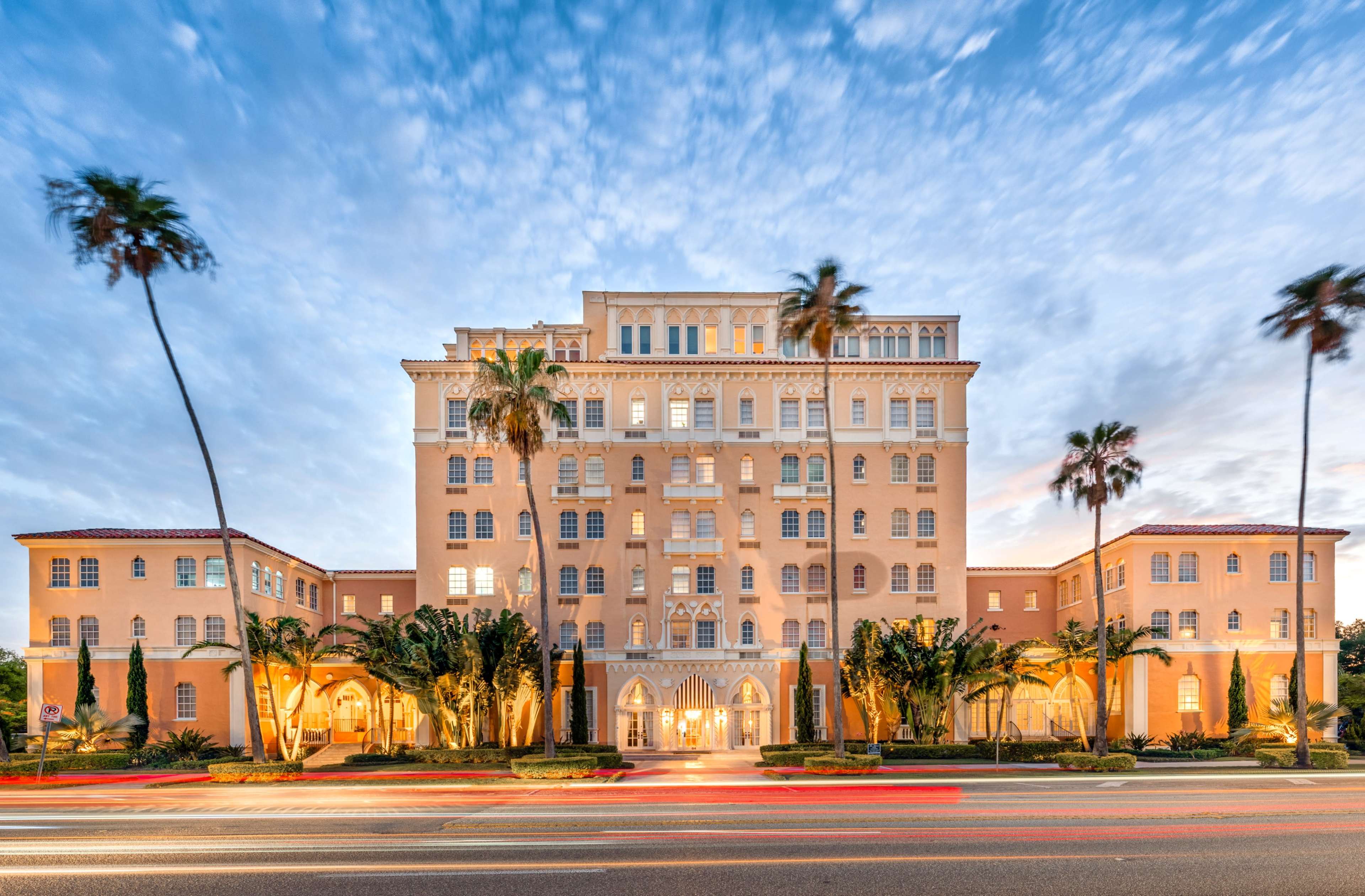 A large, pale pink hotel with multiple stories and palm trees stands against a blue sky with scattered clouds.