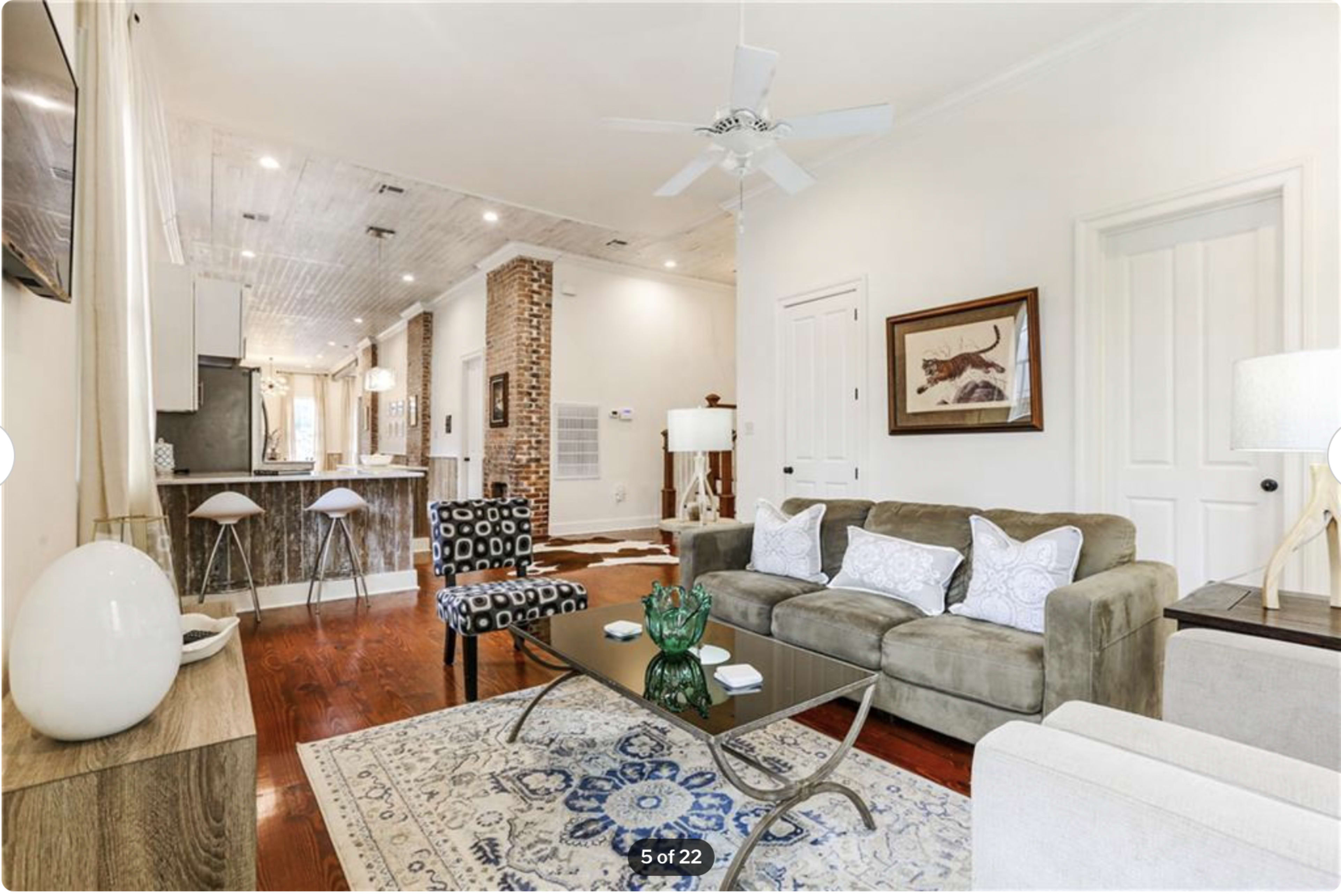 A living room with a beige sofa, patterned chairs, a coffee table, and a kitchen area in the background featuring bar stools and exposed brick.