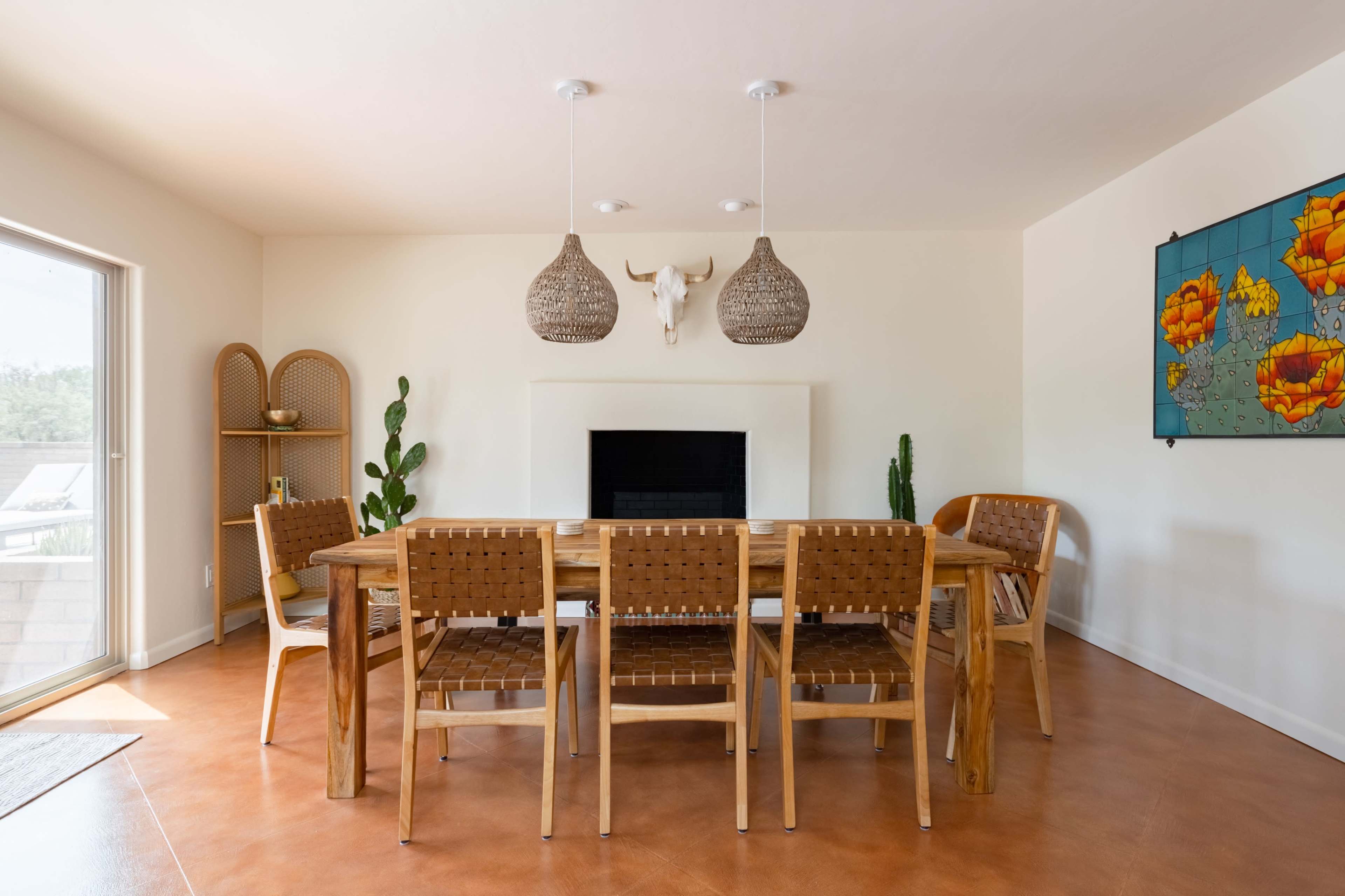 A dining area features a wooden table surrounded by chairs with woven backs, light fixtures above, and a wall adorned with a colorful artwork and decorative elements.