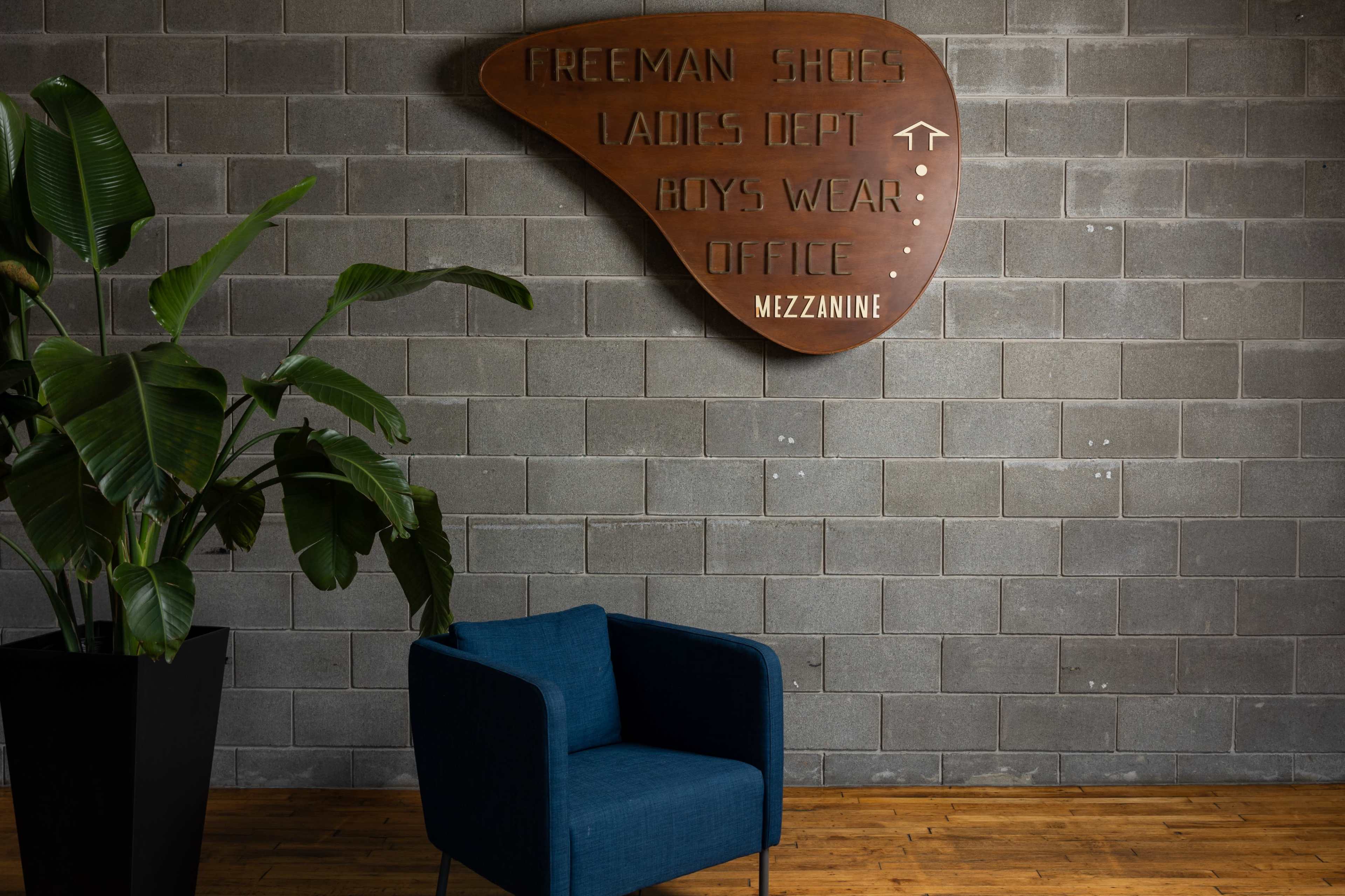 A blue armchair is positioned in front of a textured gray wall featuring a large, wooden sign with directional information about various store departments.
