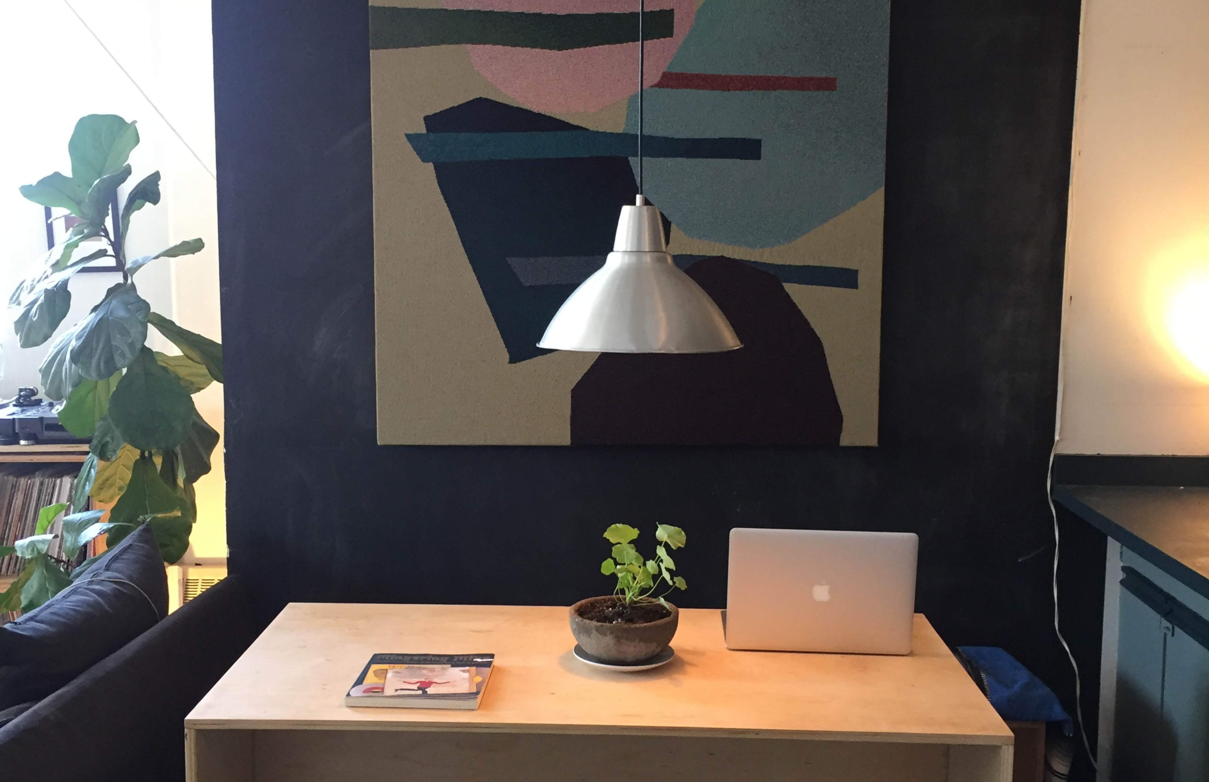 A modern workspace features a wooden desk with a laptop, a potted plant, and a large abstract artwork hanging above, complemented by a pendant light and a cozy seating area.